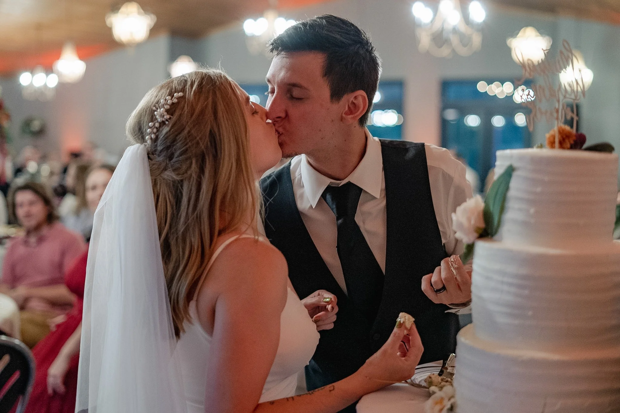 A bride and groom kissing during their wedding reception, with wedding cake visible in the foreground and guests seated in the background.