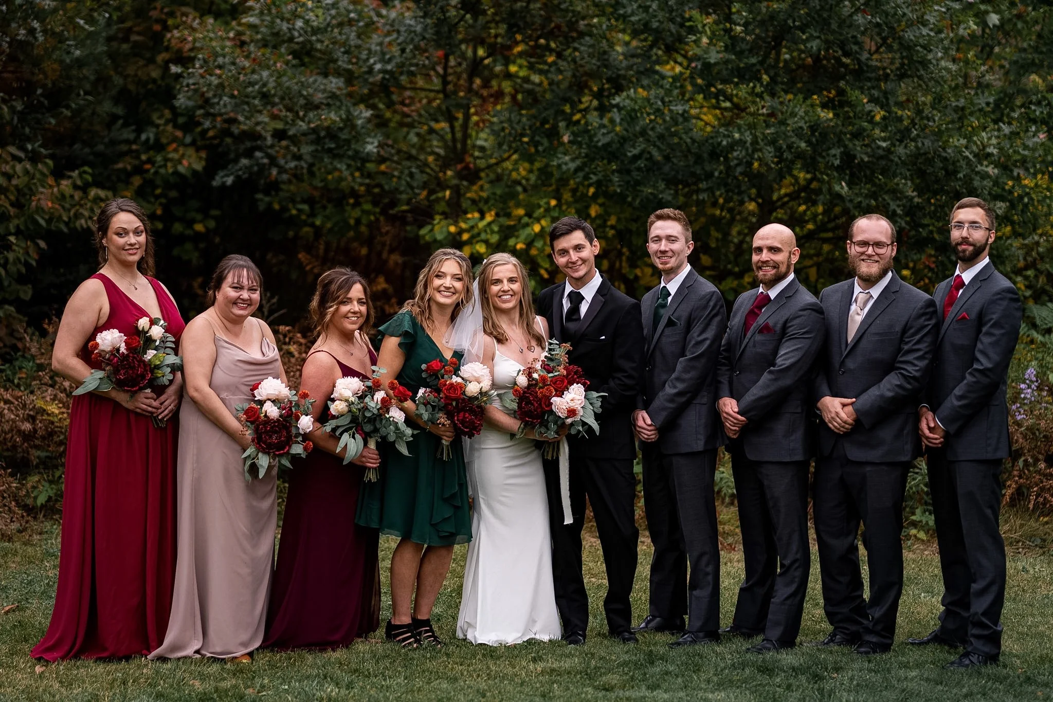 A wedding party of ten people standing outdoors in front of trees, dressed in formal attire, with the bride in a white dress and the groom in a black suit, surrounded by bridesmaids and groomsmen holding flower bouquets.