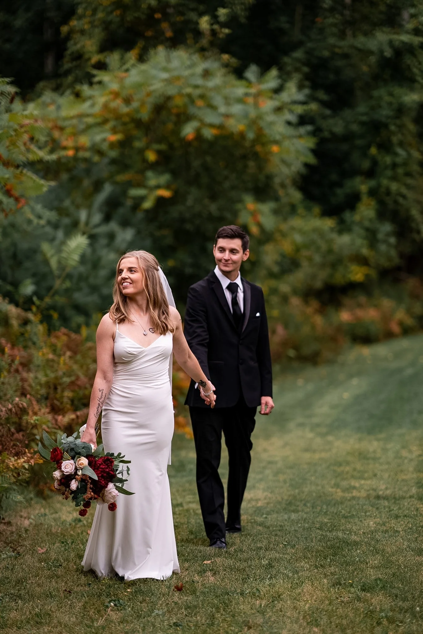A smiling bride in a white wedding gown holding a flower bouquet, walking hand-in-hand with a groom in a black suit on a grassy path surrounded by trees with green and autumn leaves.