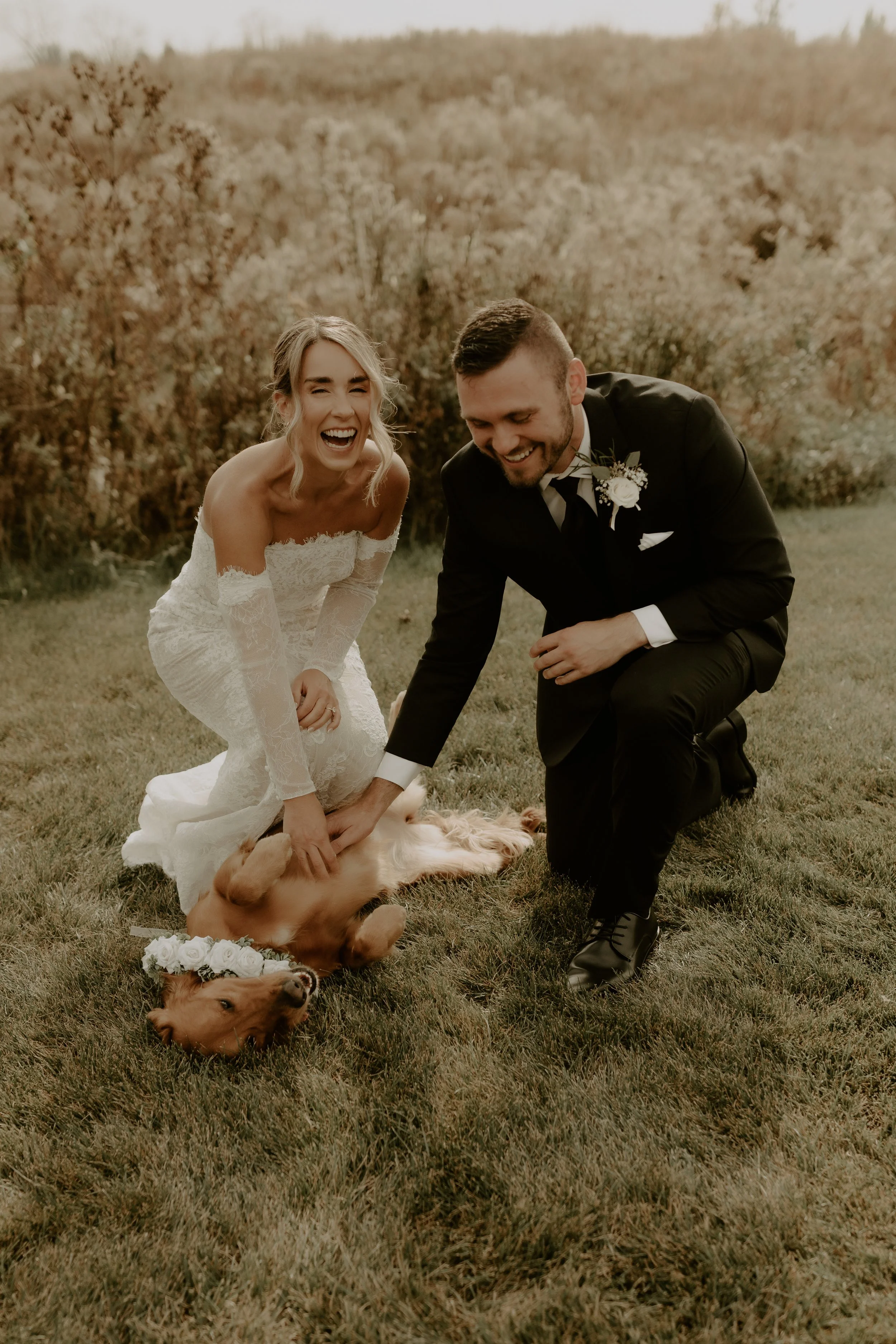 Happy bride and groom playing with a golden retriever dog lying on its back outdoors on grass, during their wedding.