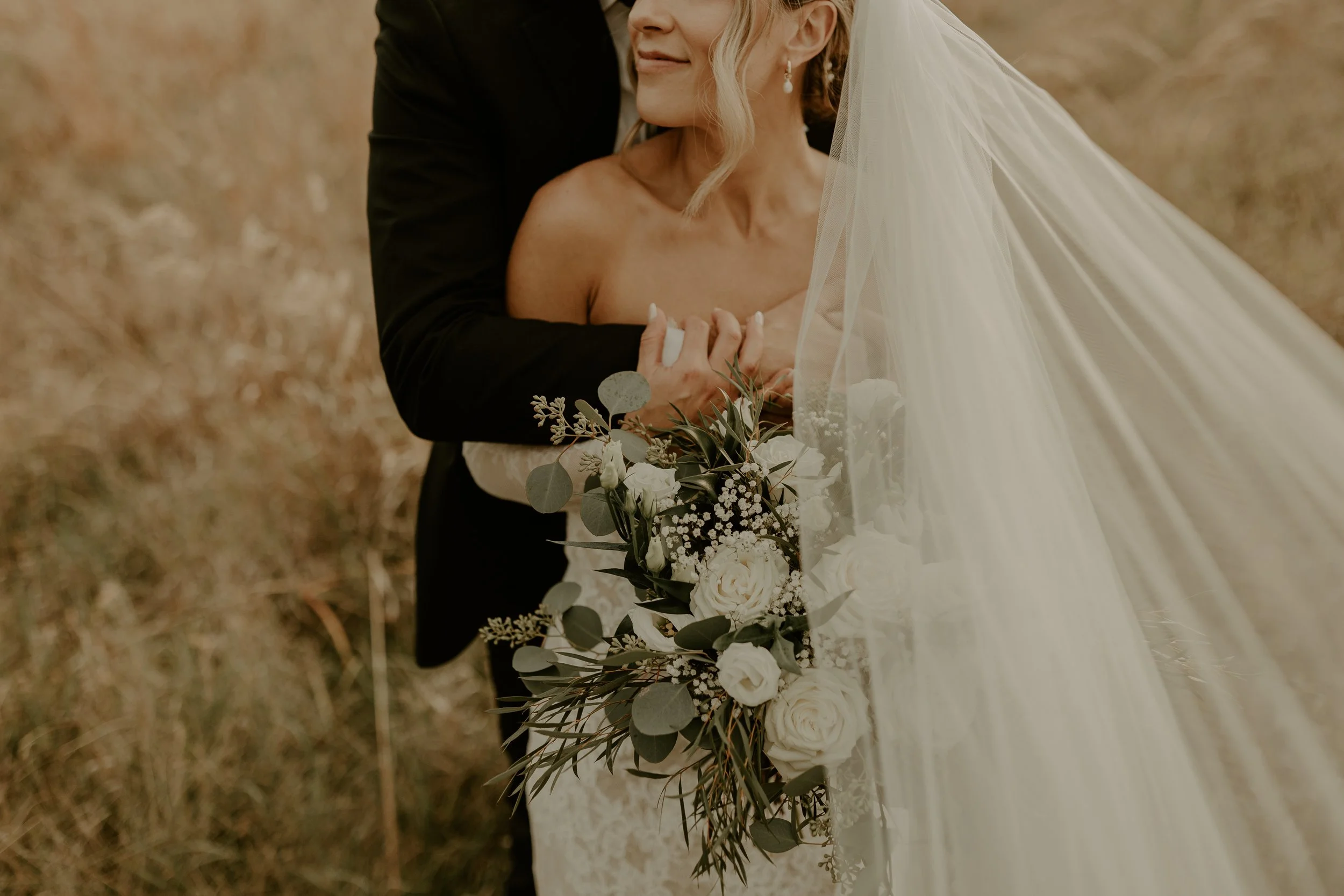 A bride and groom embracing outdoors, with the bride holding a bouquet of white roses and greenery. The bride wears a veil and earrings, and the groom is dressed in a black suit.