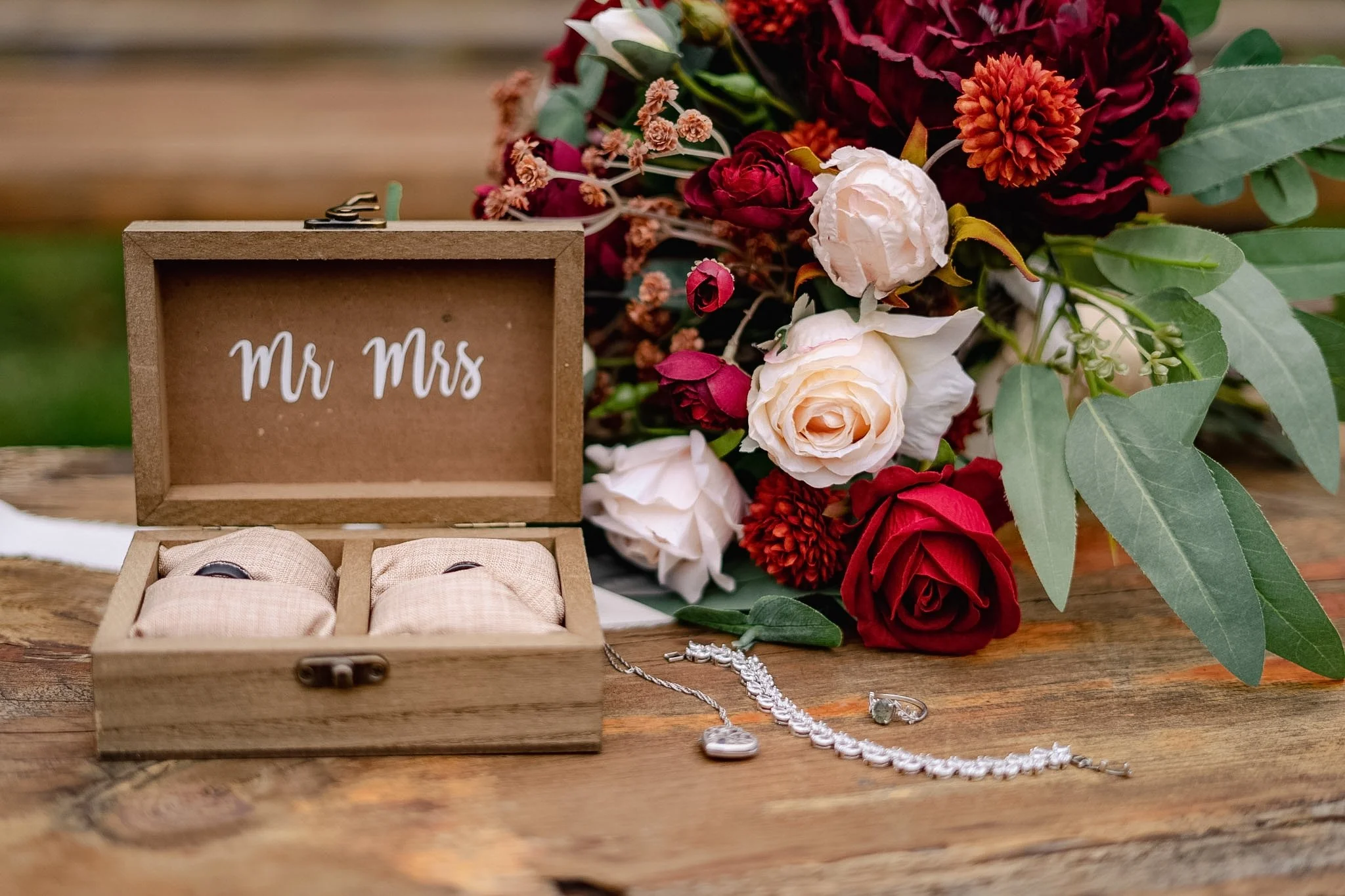 A wedding ring box with two rings beside a bridal necklace and a ring on a wooden table, with a bouquet of red, white, and orange flowers in the background.