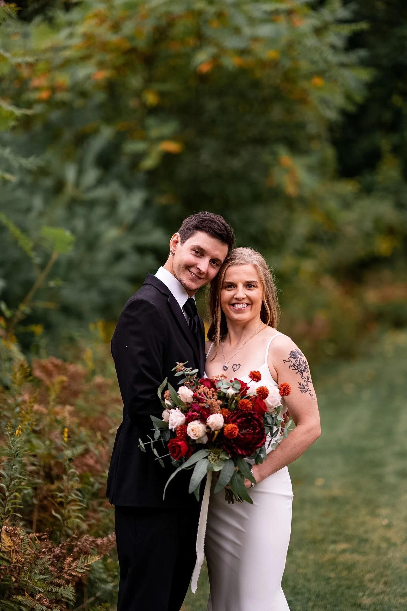 A newlywed couple standing outdoors on a grassy path, smiling, with the bride holding a large bouquet of red, pink, and orange flowers, surrounded by green foliage and trees.
