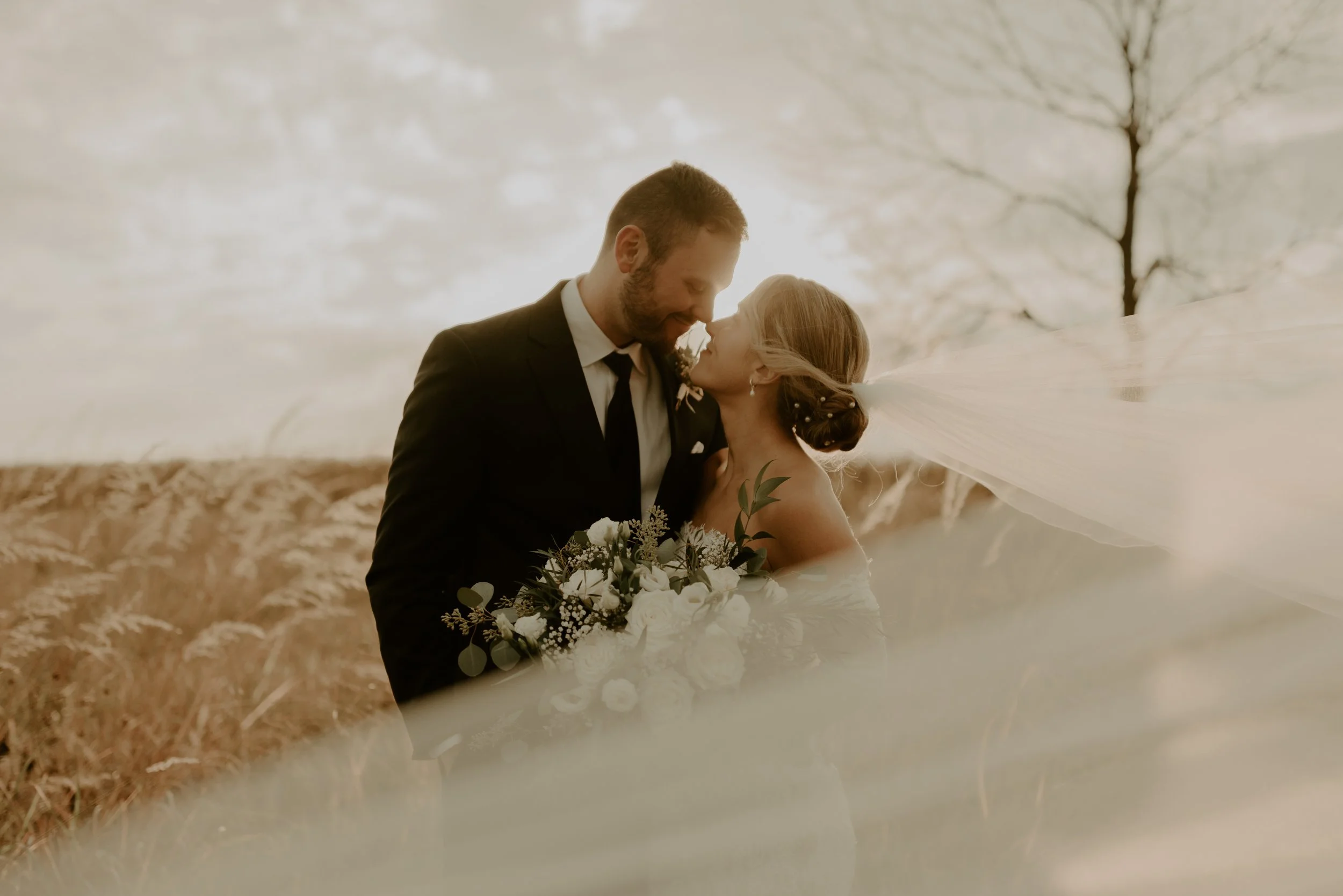 A bride and groom sharing a romantic moment outdoors, with the bride holding a bouquet and her veil flowing in the wind against a background of autumn landscape and bare trees.