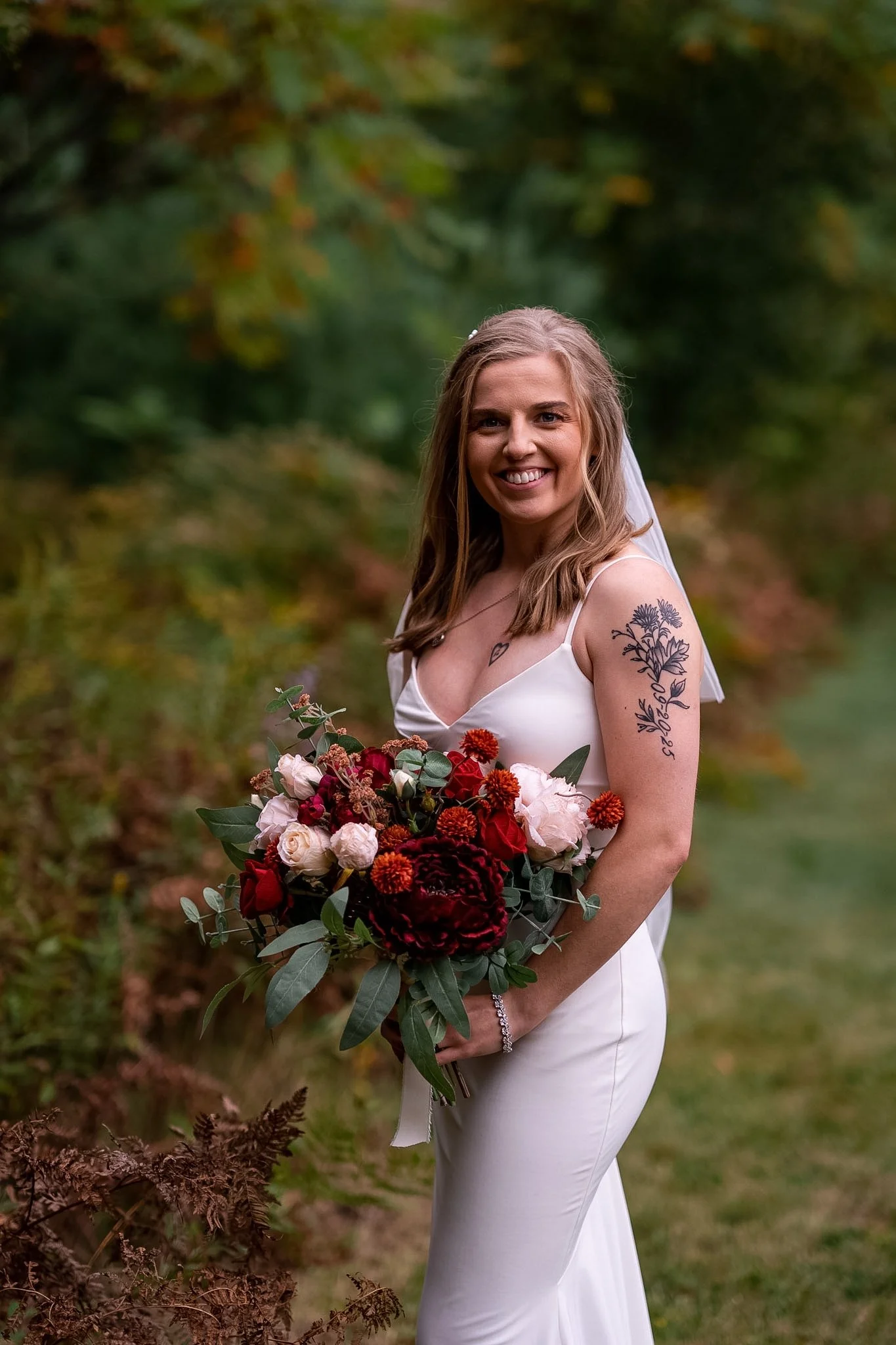 A smiling bride in a white wedding dress holding a large bouquet of red, white, and pink flowers outdoors, with a backdrop of green foliage.