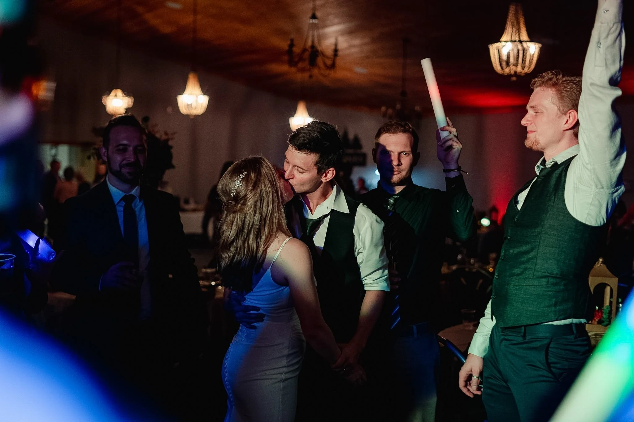 A couple kisses at a wedding reception surrounded by friends. One man is holding a glow stick and dancing. The background has decorative chandeliers and warm lighting.