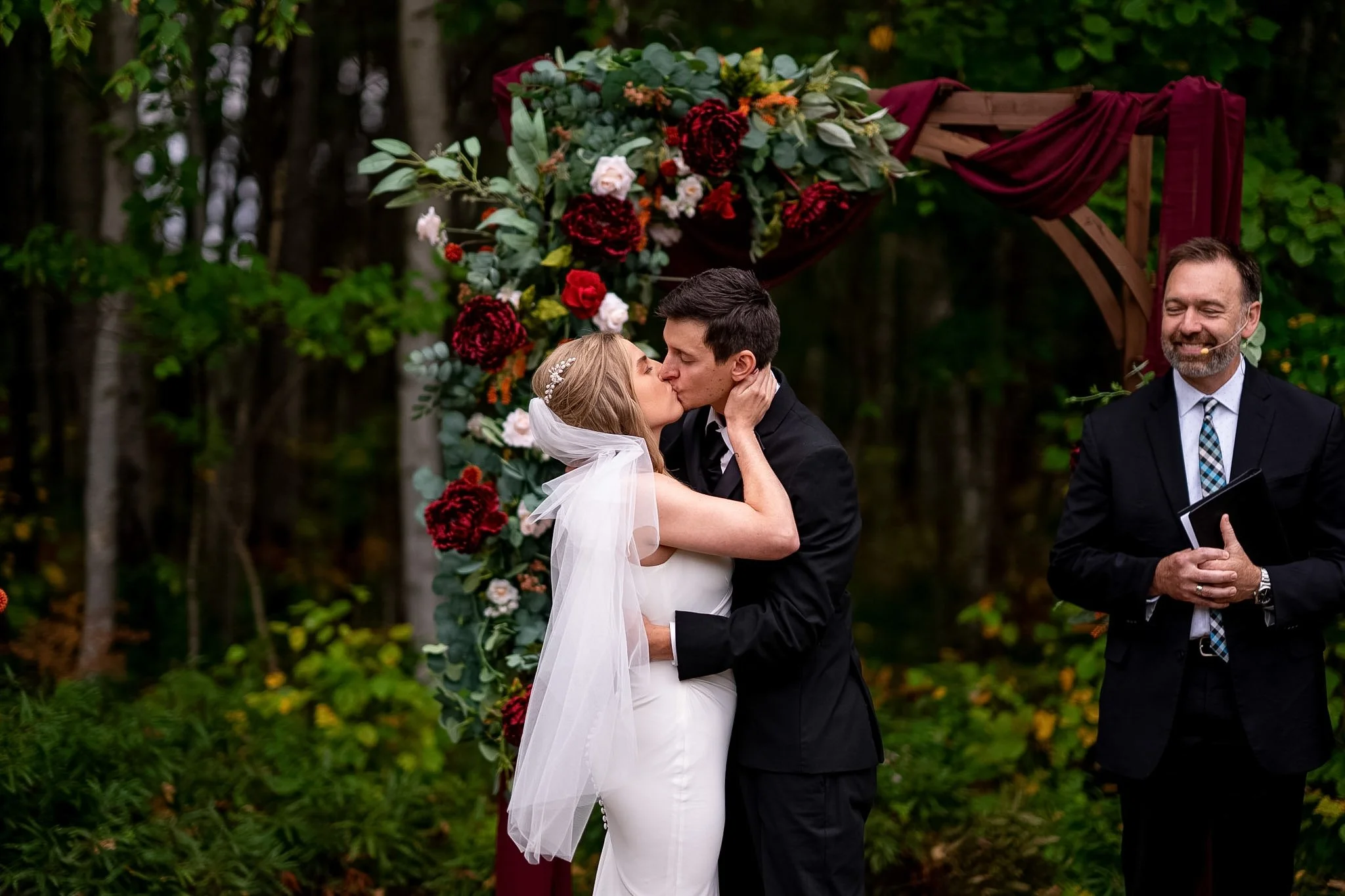 A bride and groom share a kiss during their outdoor wedding ceremony, with a floral arch behind them and a officiant standing nearby, all in a forest setting.