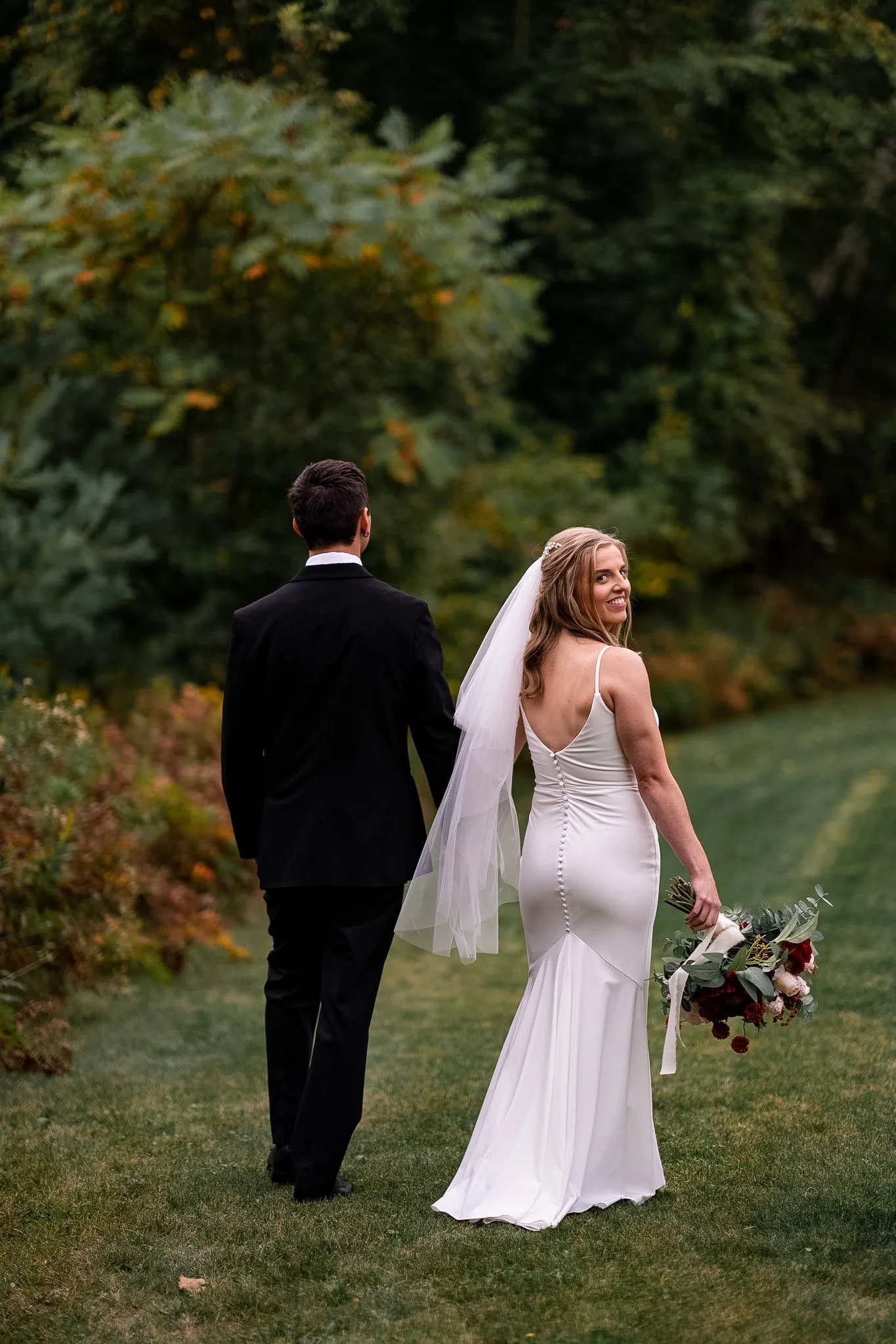 Bride in a white gown holding a bouquet, walking hand-in-hand with groom in a black suit outdoors surrounded by greenery.