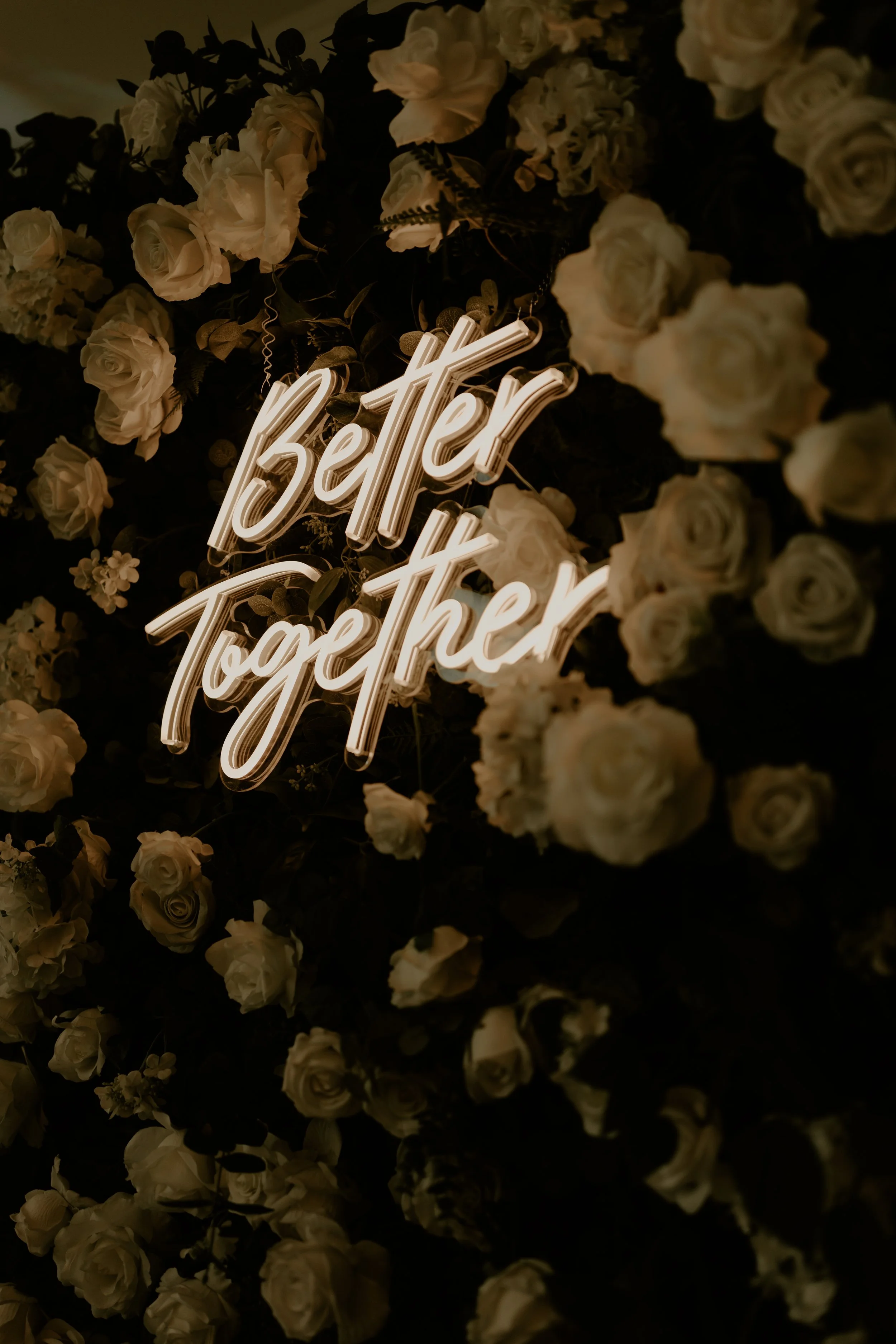 A neon sign that says 'better together' surrounded by white roses and dark greenery.