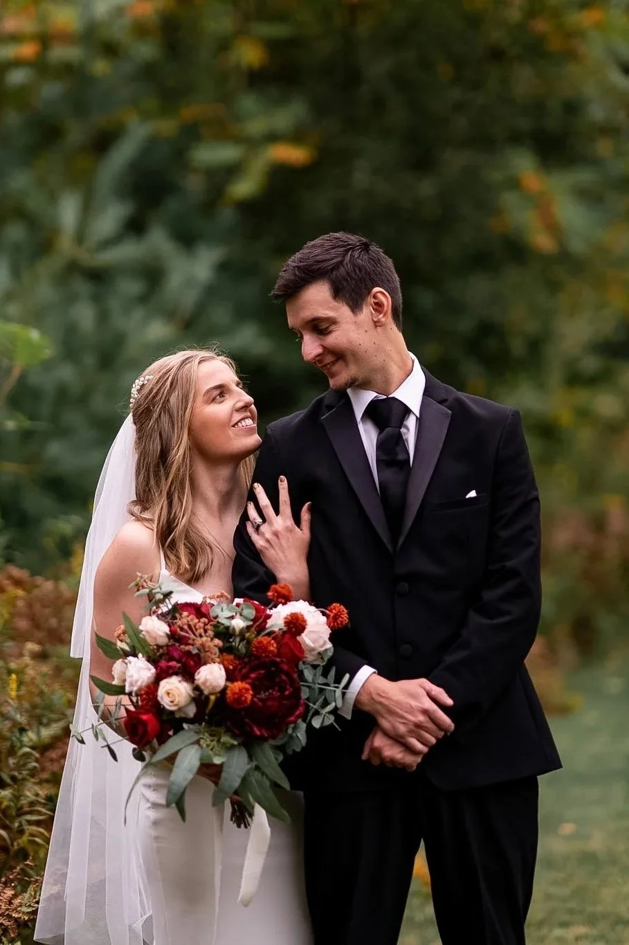 A bride and groom are standing outdoors in a garden, sharing a loving moment. The bride holds a large bouquet of red, white, and pink flowers, and they are gazing at each other tenderly.