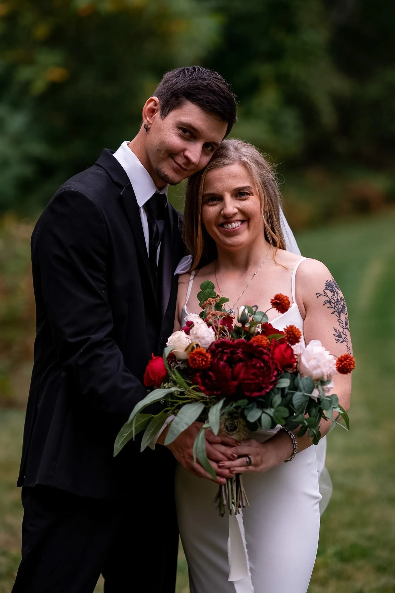 A newlywed couple smiling outdoors, with the groom in a black suit and the bride in a white dress holding a large bouquet of red, white, and pink flowers.