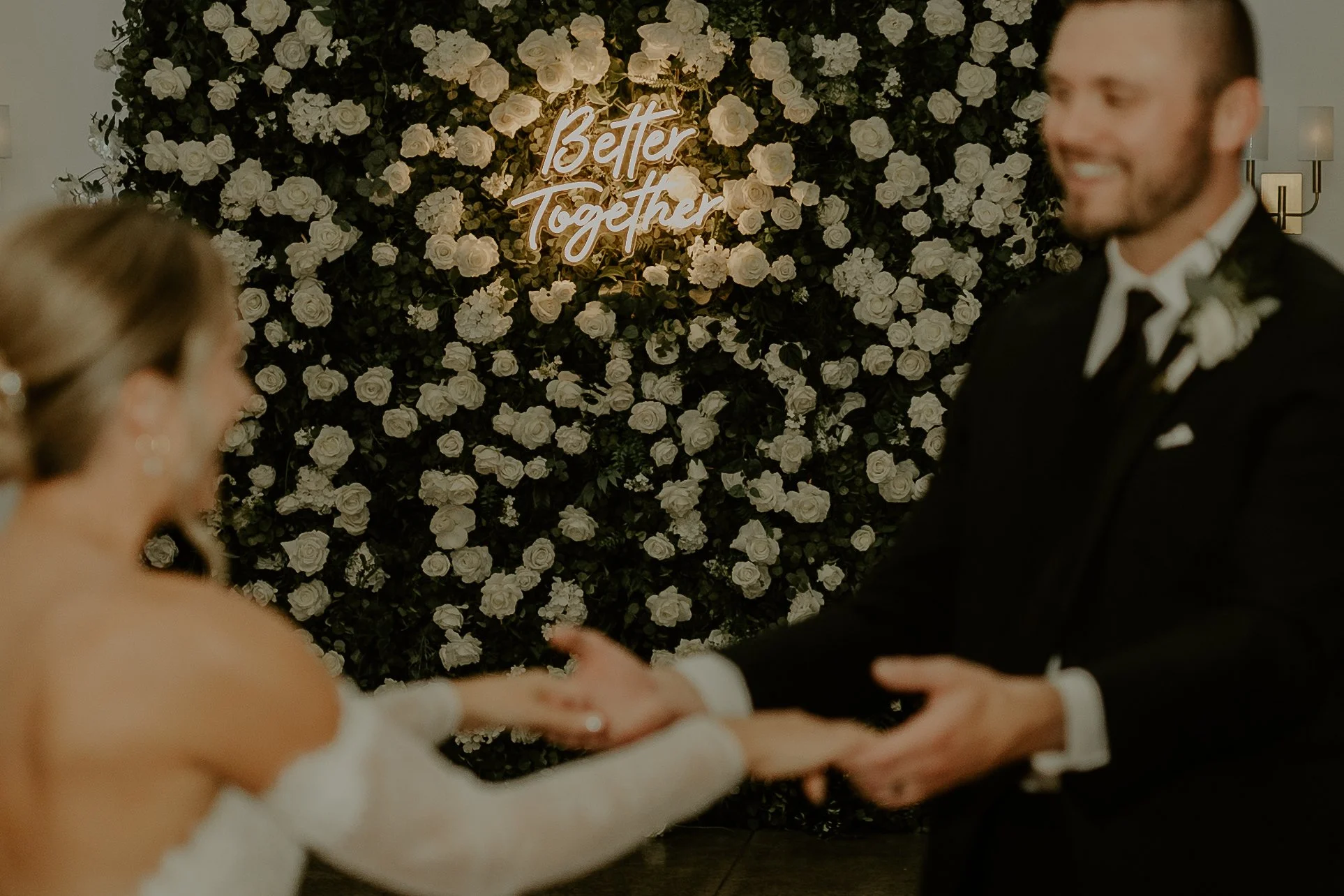A bride and groom holding hands during their wedding ceremony, with a background of white roses and a neon sign that reads 'Better Together'.
