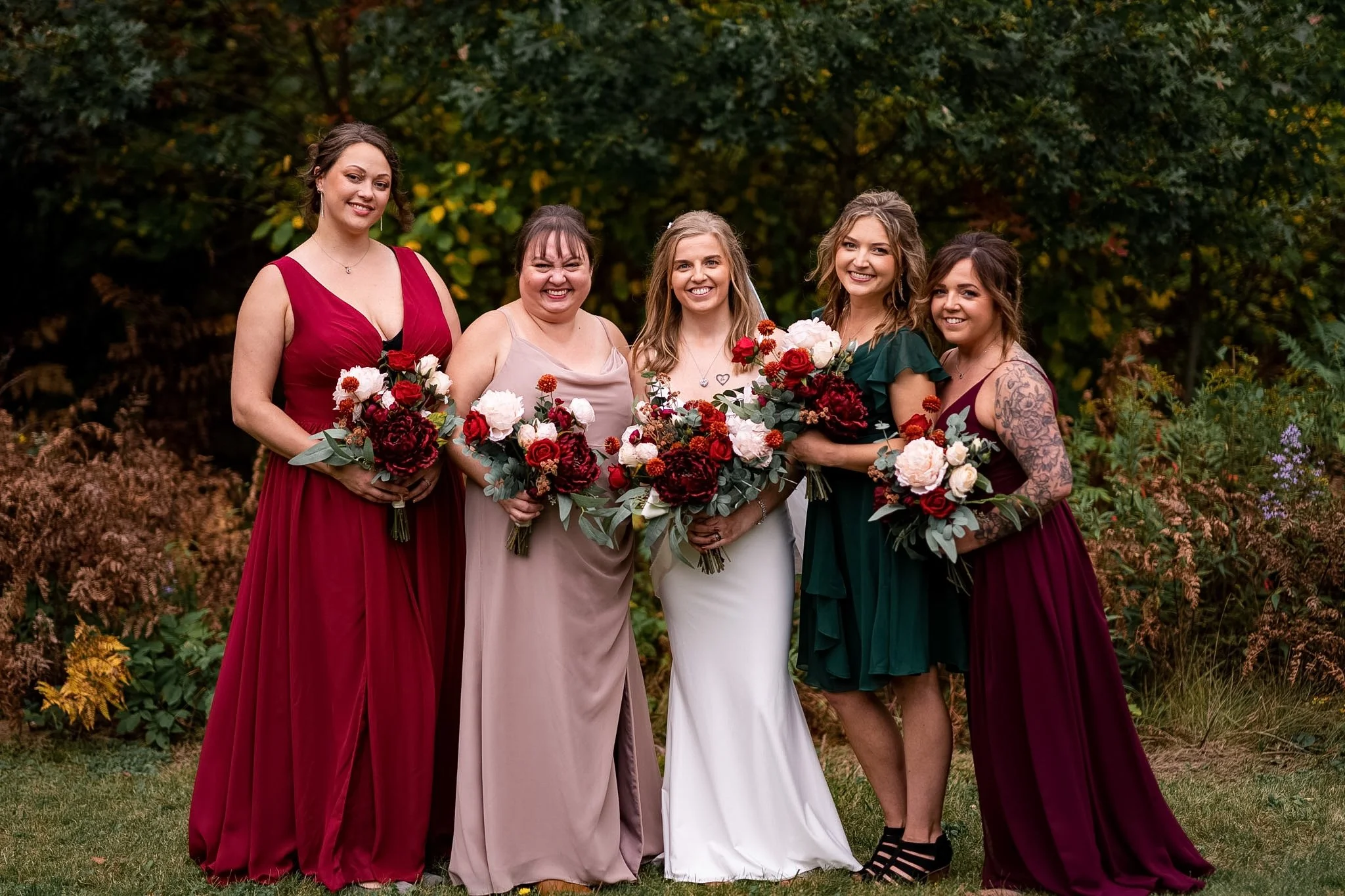 Group of five women in colorful dresses holding bouquets of flowers, standing outdoors with trees in the background.