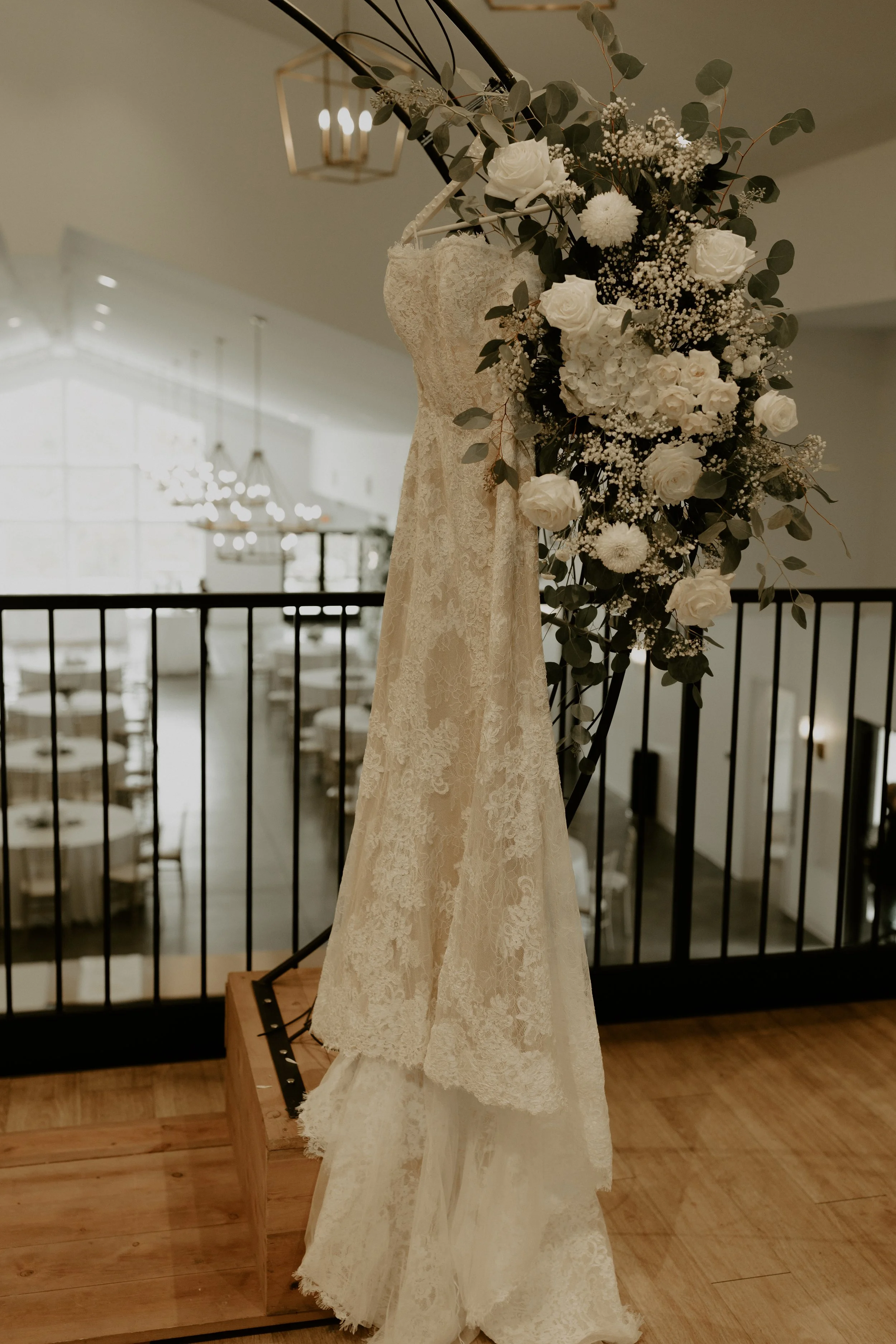 A wedding dress on a mannequin adorned with flowers and greenery, displayed on a wooden platform in a well-lit indoor setting.
