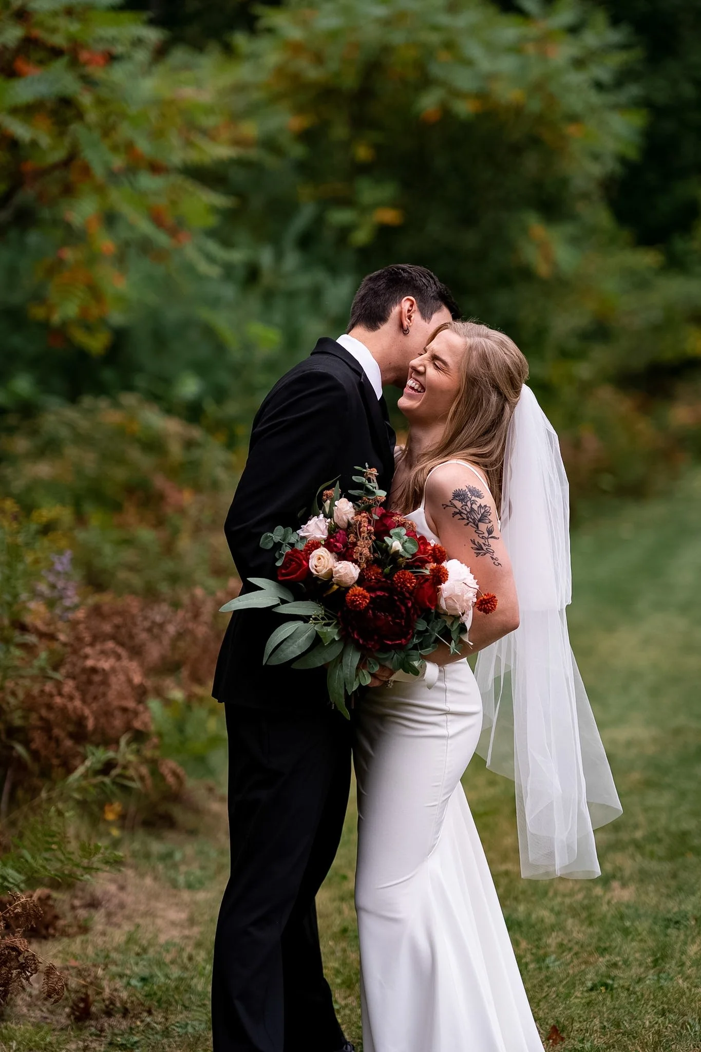 A newlywed couple in wedding attire sharing a joyful moment outdoors, with the groom whispering or kissing the bride on the forehead, surrounded by greenery and colorful foliage.