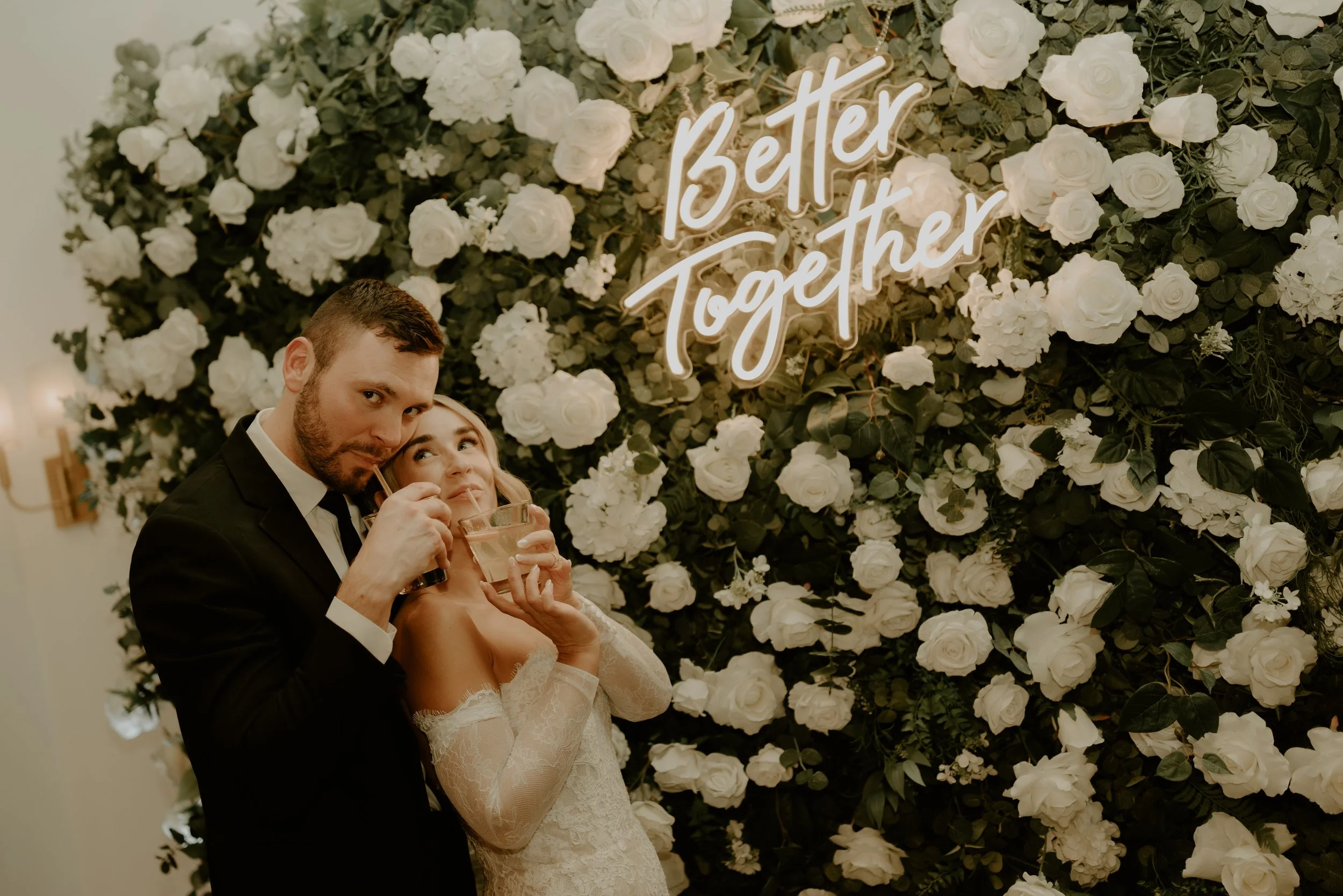 A bride and groom posing together at a wedding reception, standing in front of a floral wall with white flowers and green leaves, with a neon sign that reads 'Better Together.'