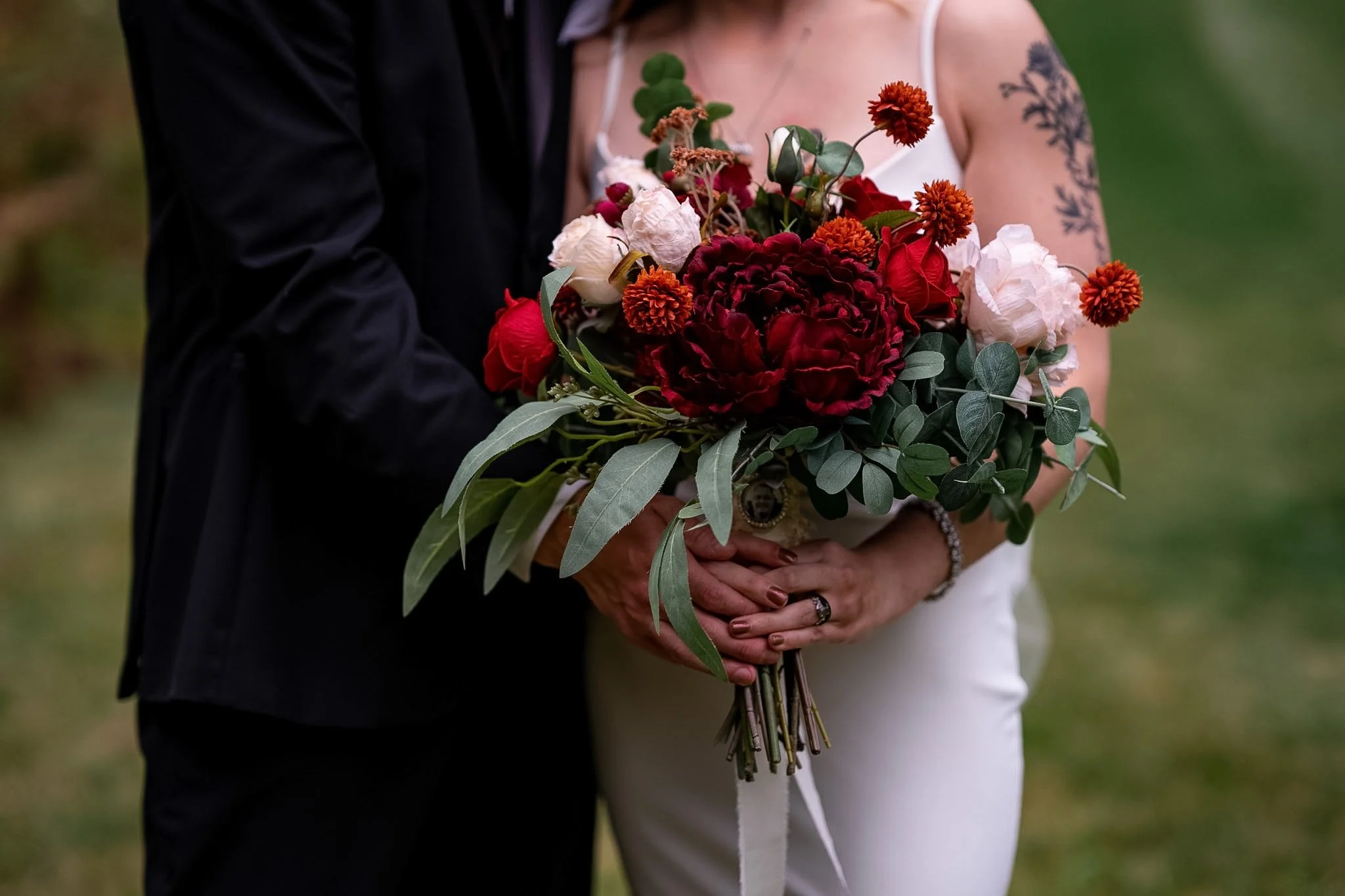 A bride and groom holding a bouquet of red, white, and orange flowers with green foliage, outdoors.
