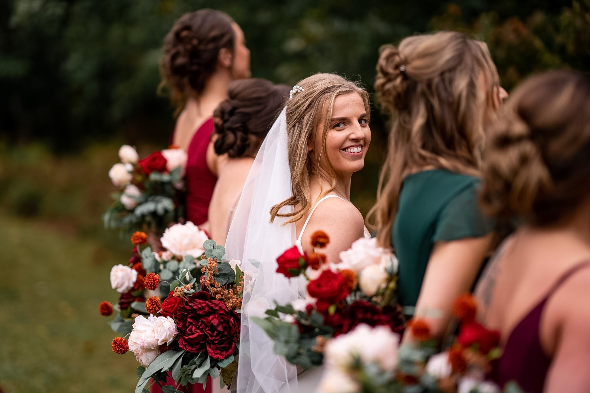 A bride and bridesmaids at a wedding outdoors, with the bride smiling and looking at the camera, holding bouquets of red, pink, and white flowers.