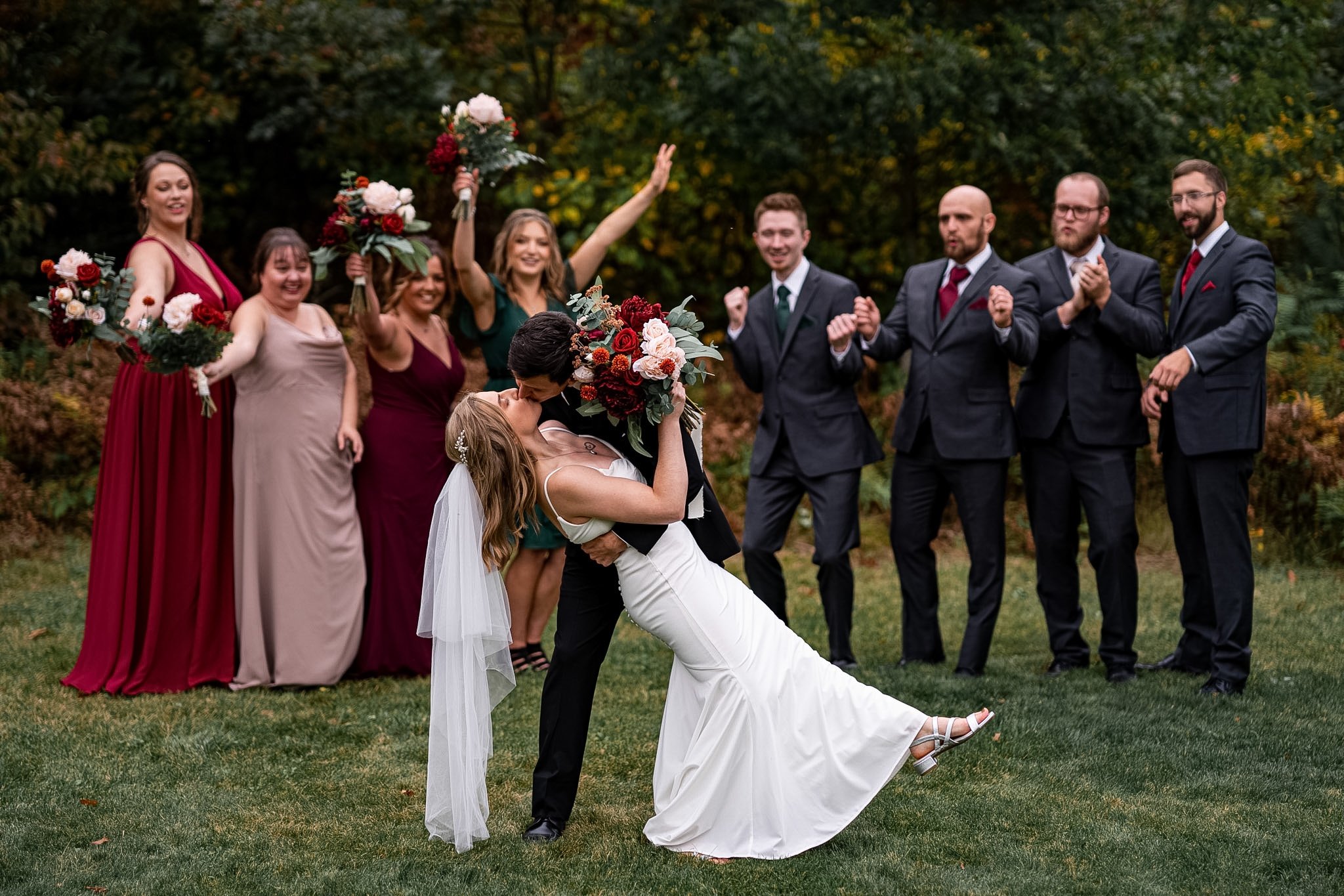 A newlywed couple sharing a kiss, with the groom dipping the bride, who is holding a large bouquet of flowers. Bridesmaids and groomsmen cheer in the background on a grassy outdoor area surrounded by trees.