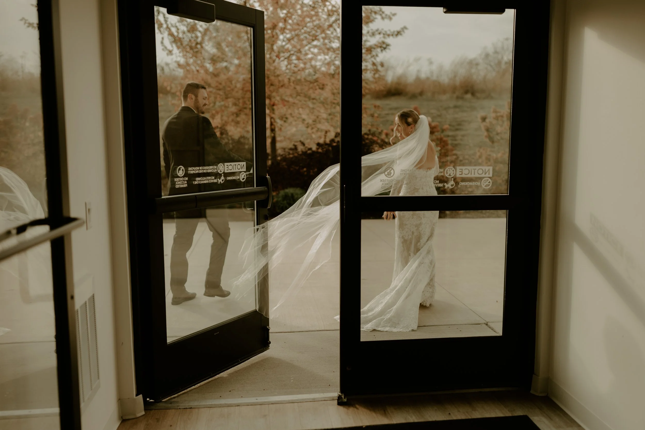 A bride and groom standing outside a glass entrance door on their wedding day, with the bride wearing a long white lace gown and veil, and the groom in a black suit, with autumn trees visible in the background.