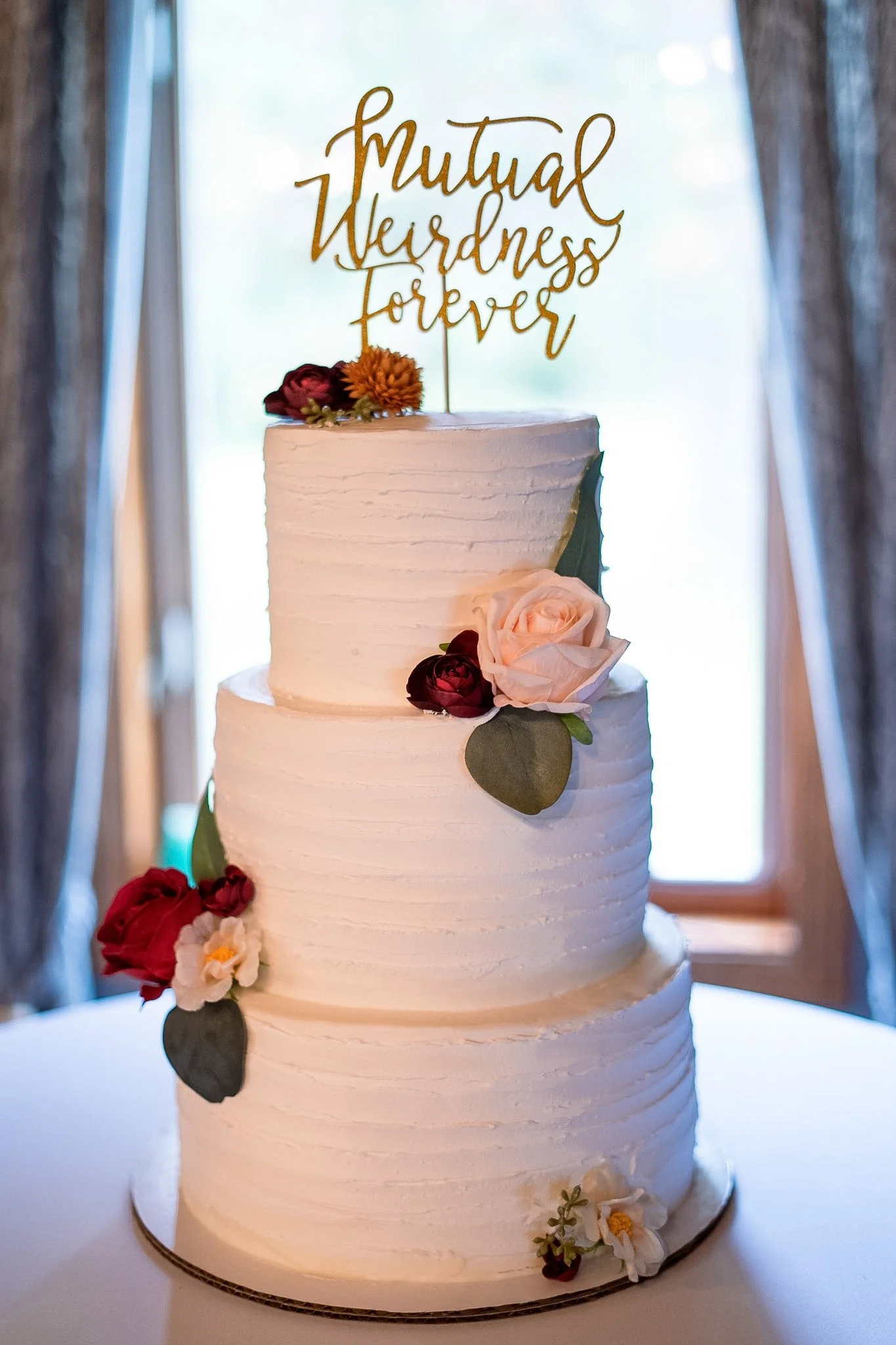 Three-tier white wedding cake with flowers and a gold topper saying 'mutual weirdness forever', placed on a table with a blurred window background.