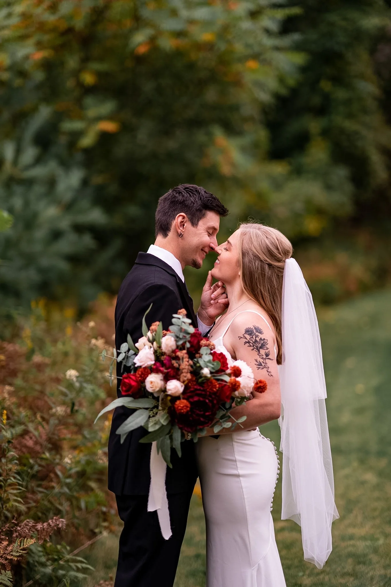 A newlywed couple on their wedding day, standing close outdoors with a lush green background. The groom, dressed in a black suit, and the bride, in a white gown with a veil, are smiling and touching noses. The bride holds a large bouquet of red, pink