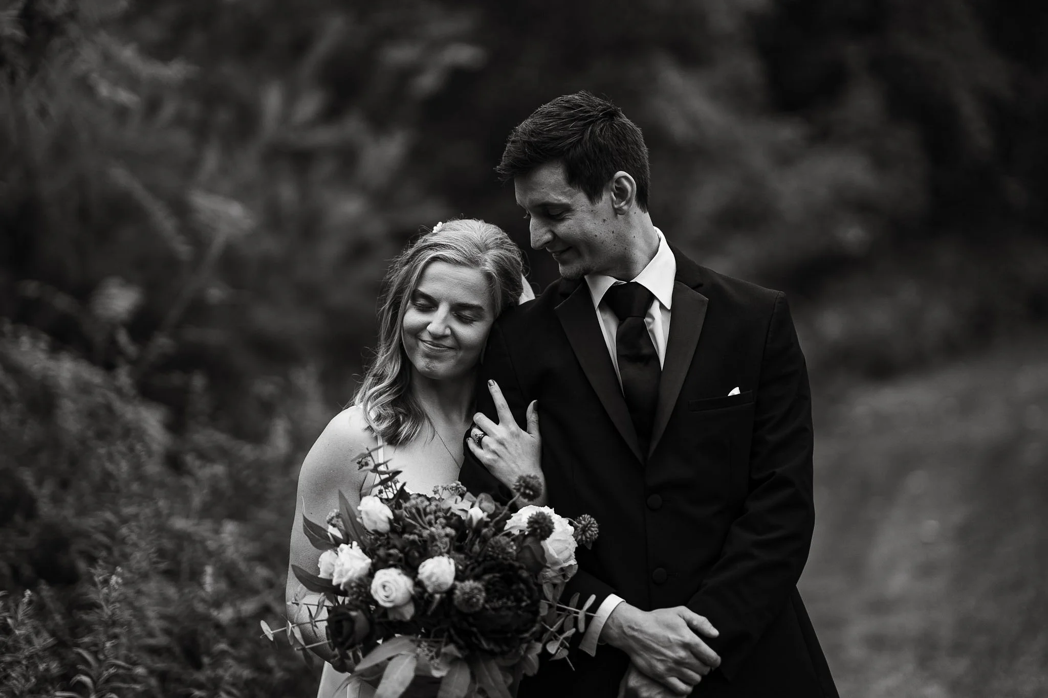 Black and white photo of a bride and groom standing closely outdoors, smiling gently. The bride holds a bouquet of flowers, and the groom has his hand on her arm. They are dressed in wedding attire, surrounded by natural scenery.