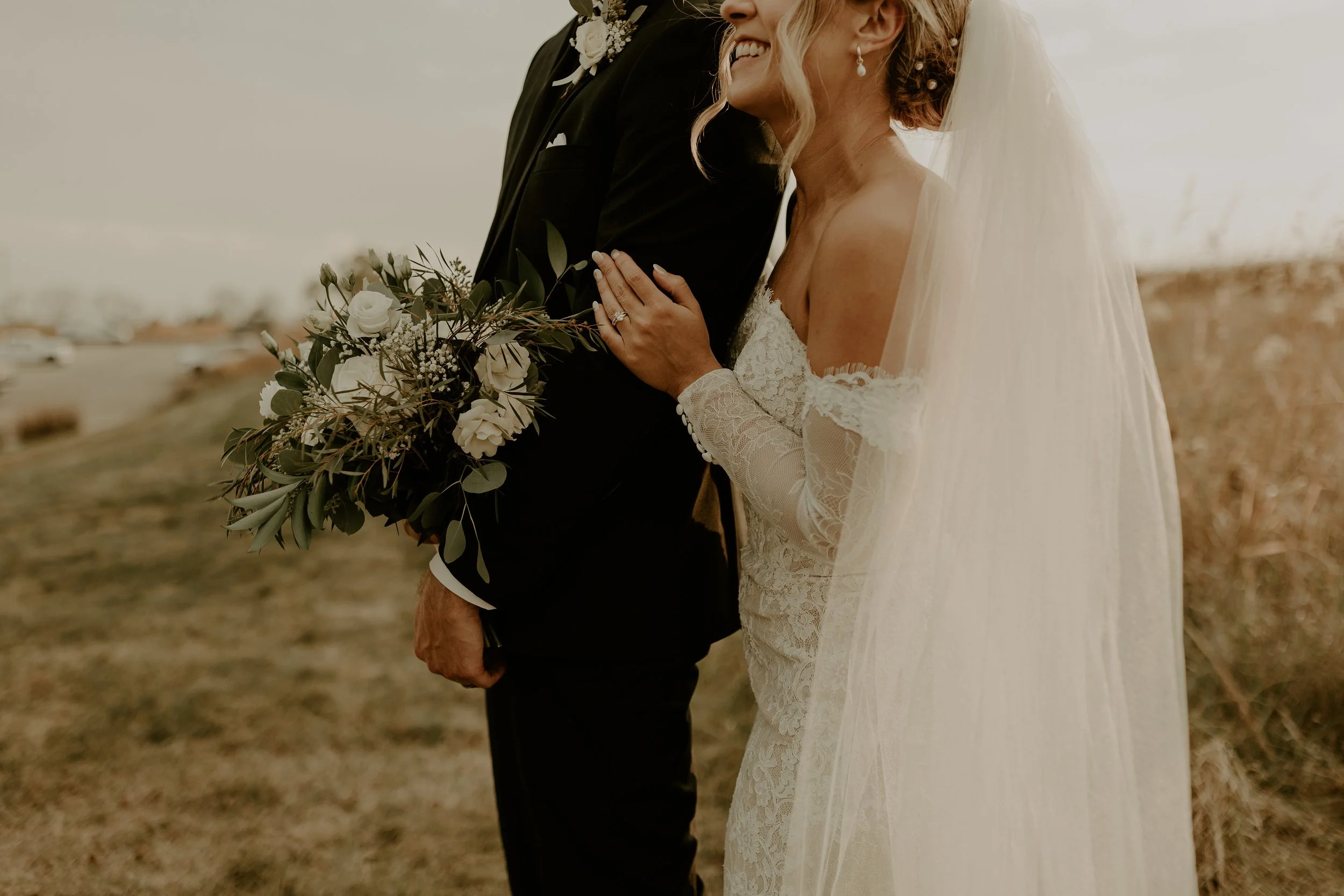 Close-up of a bride and groom on their wedding day outdoors, with the bride's hand resting on the groom's chest and holding a bouquet of white flowers and greenery.