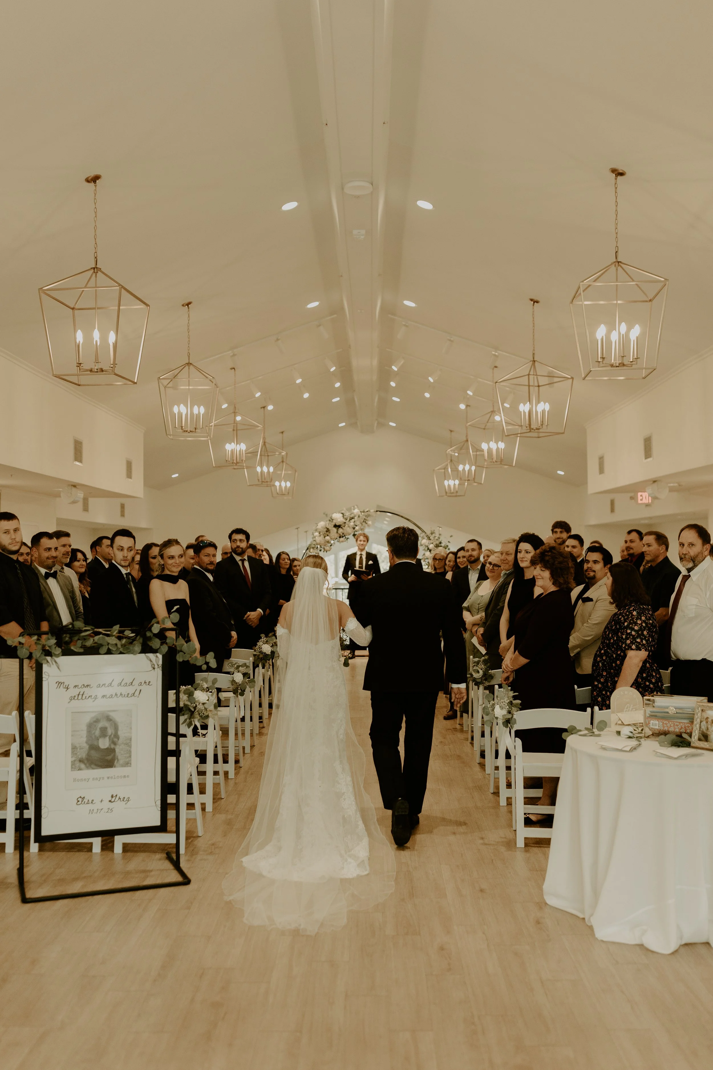 A bride and groom walking down the aisle at their wedding ceremony in a spacious hall, with guests standing on both sides, decorated with white flowers and greenery.