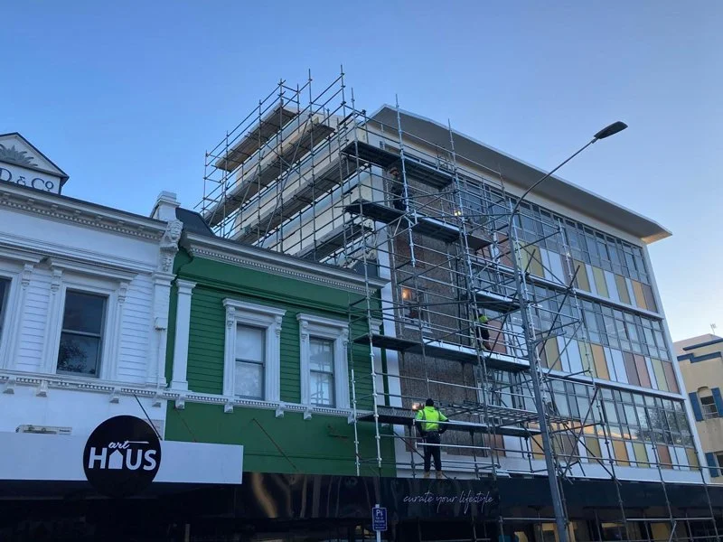 Construction worker in a high-visibility vest on scaffolding in front of a green and white building on a city street.
