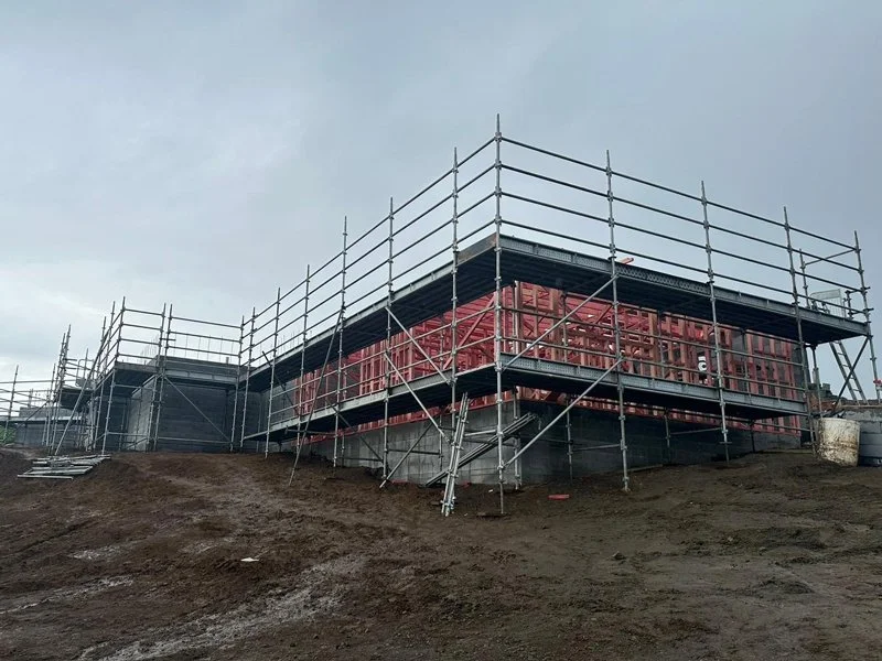 Construction site with a building under construction surrounded by metal scaffolding, on a muddy ground under a cloudy sky.
