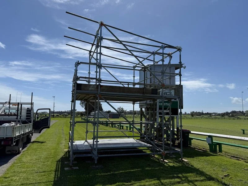 A metal scaffolding structure set up on a grassy field near a sports track, with trucks parked nearby and an open sky above.