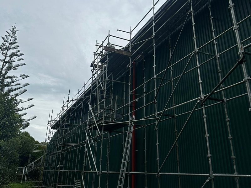 Construction scaffolding set up along a large green industrial building with a ladder leaning against it, under a cloudy sky.