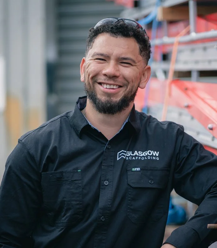 Smiling man wearing a black shirt with a 'Glasgow Scaffolding' logo, standing in front of scaffolding equipment.