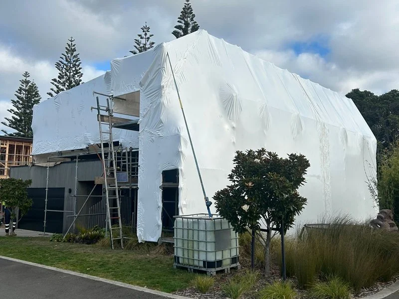 A house under construction or renovation, wrapped in white construction material with scaffolding and ladder outside, surrounded by trees and plants.