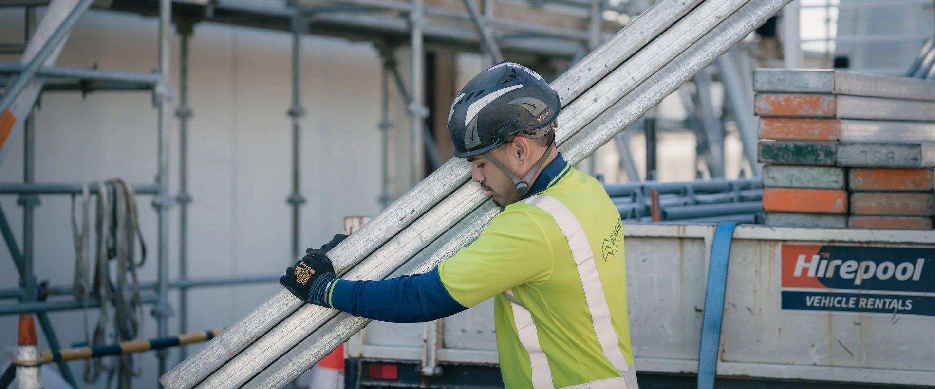Construction worker wearing a helmet and high-visibility vest carrying metal pipes on a construction site.