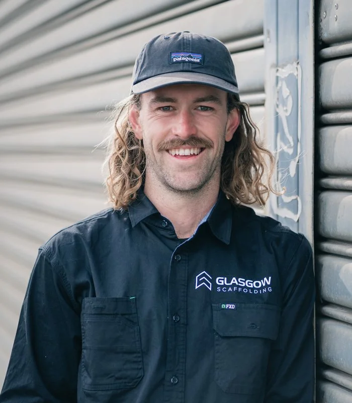 A man with long curly blonde hair and a mustache smiling, wearing a dark Patagonia cap and a black shirt with Glasgow Scaffolding logo, standing next to a gray metal rolling shutter.