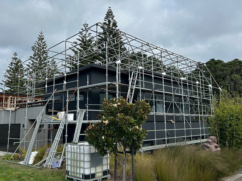 A modern, dark-colored house under construction with metal scaffolding around it, surrounded by trees and grass.