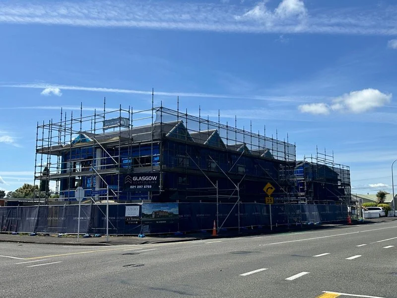 A building under construction surrounded by blue scaffolding and safety barriers on a street with cars and traffic signs under a blue sky.
