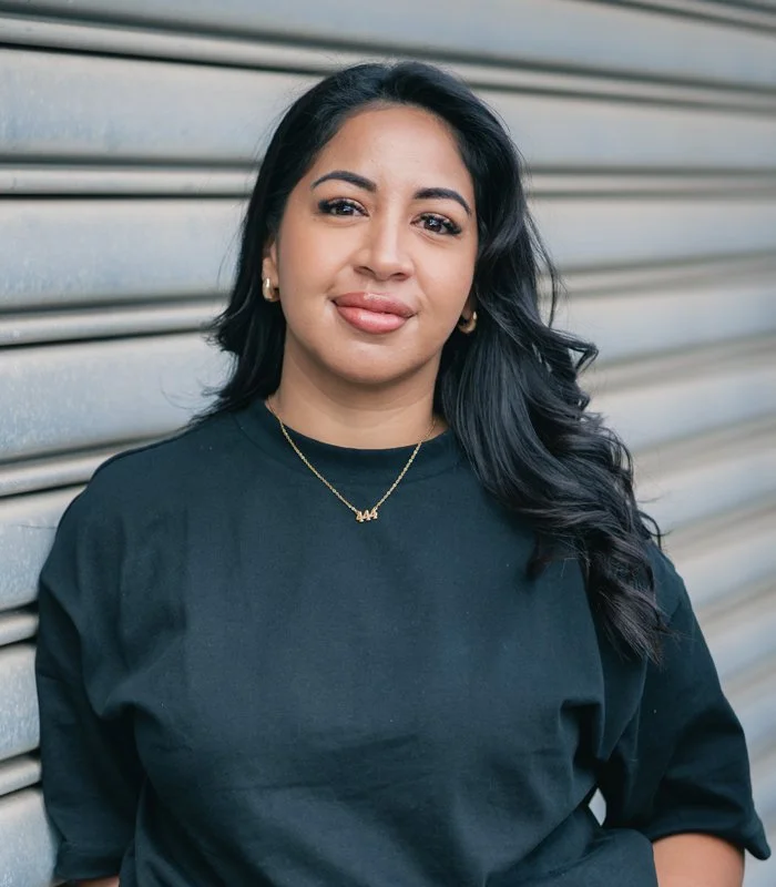 A woman with long black hair, wearing a black shirt and gold necklace, standing against a metallic garage door.