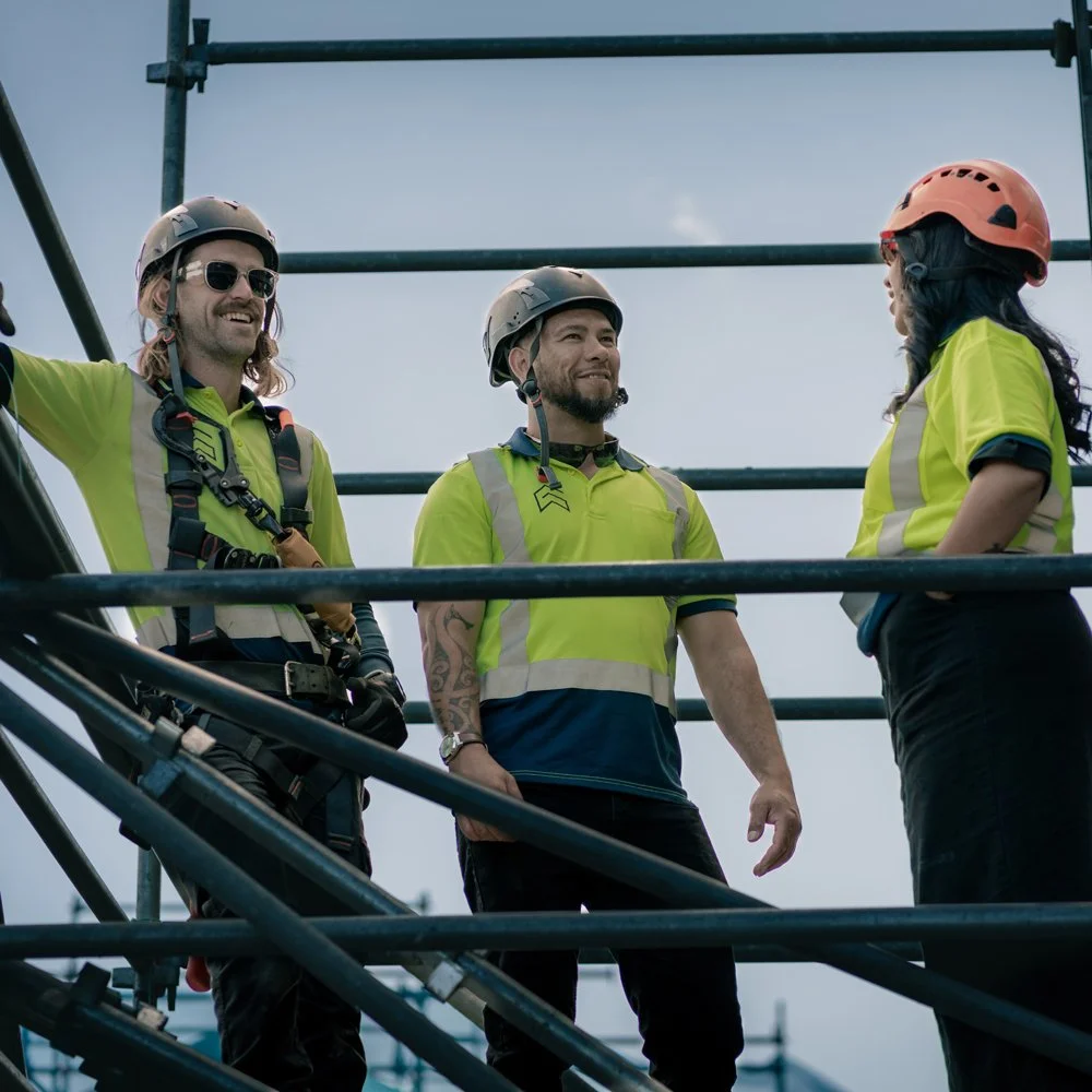 Three Glasgow Scaffolding workers wearing high-visibility uniforms and safety helmets and high-visibility shirts talking on scaffolding at a construction site.