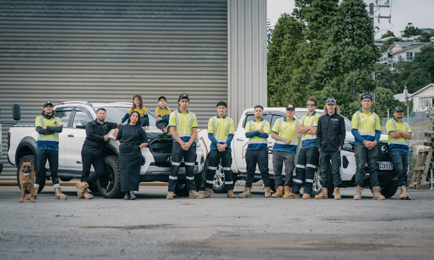 Group of people, including children and adults, standing in front of vehicles, wearing high-visibility clothing and work boots, with a dog present, outdoors near a metal building.