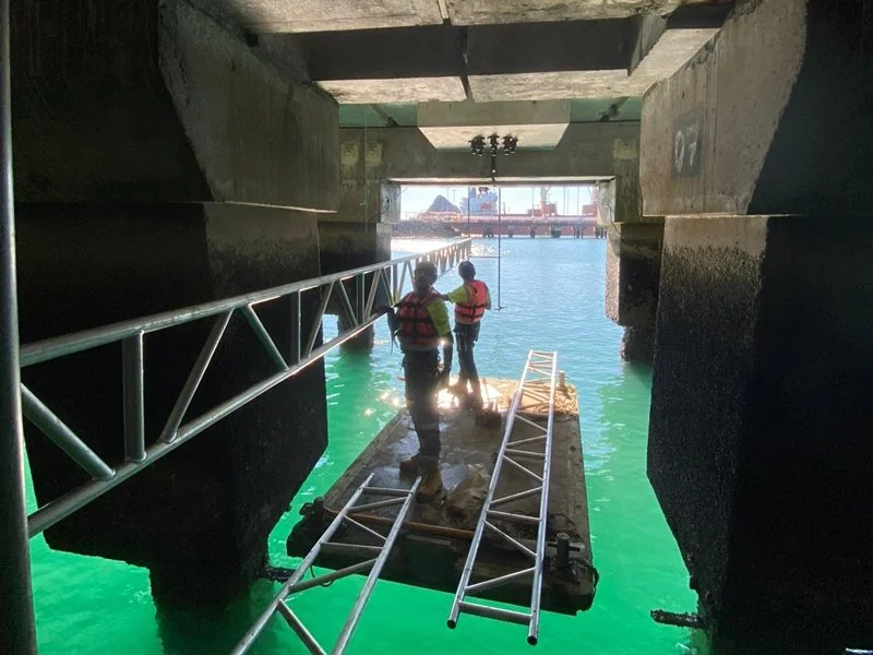 Two workers on a floating platform under a bridge, with ladders and safety gear, working near the water.