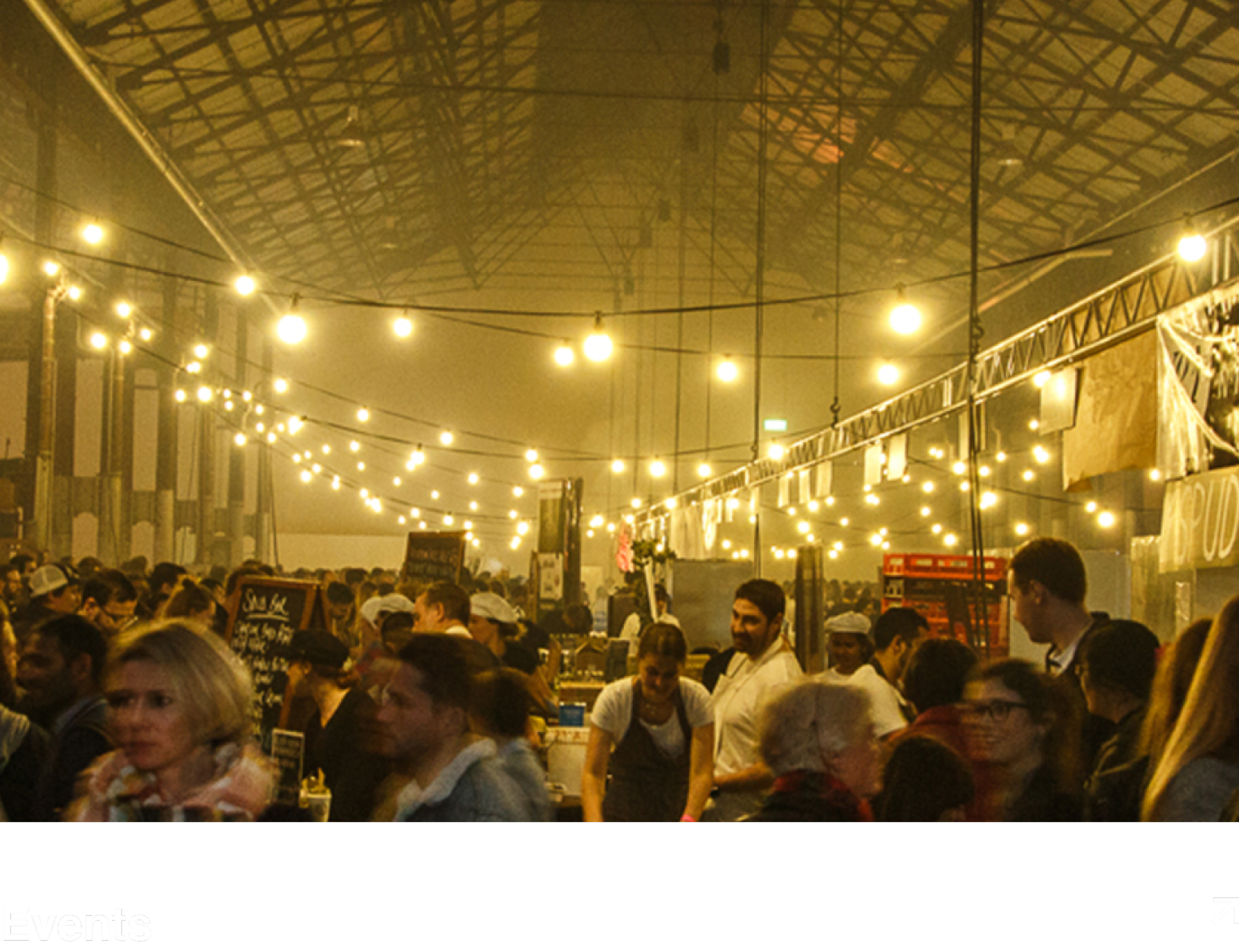 A bustling indoor event space with strings of warm yellow lights hanging overhead. Crowds of people, including vendors and attendees, are gathered, some in white uniforms, indicating possibly a market, fair, or festival.