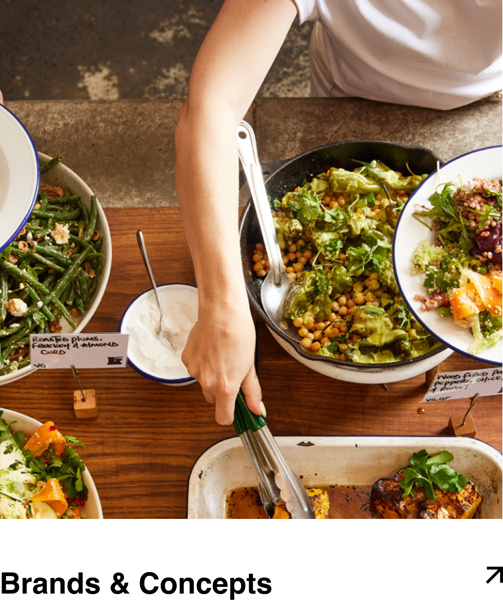 A person serving food from a buffet table with various salads and dishes. The table includes green beans, chickpea salad, roasted tofu, and other vegetable dishes.