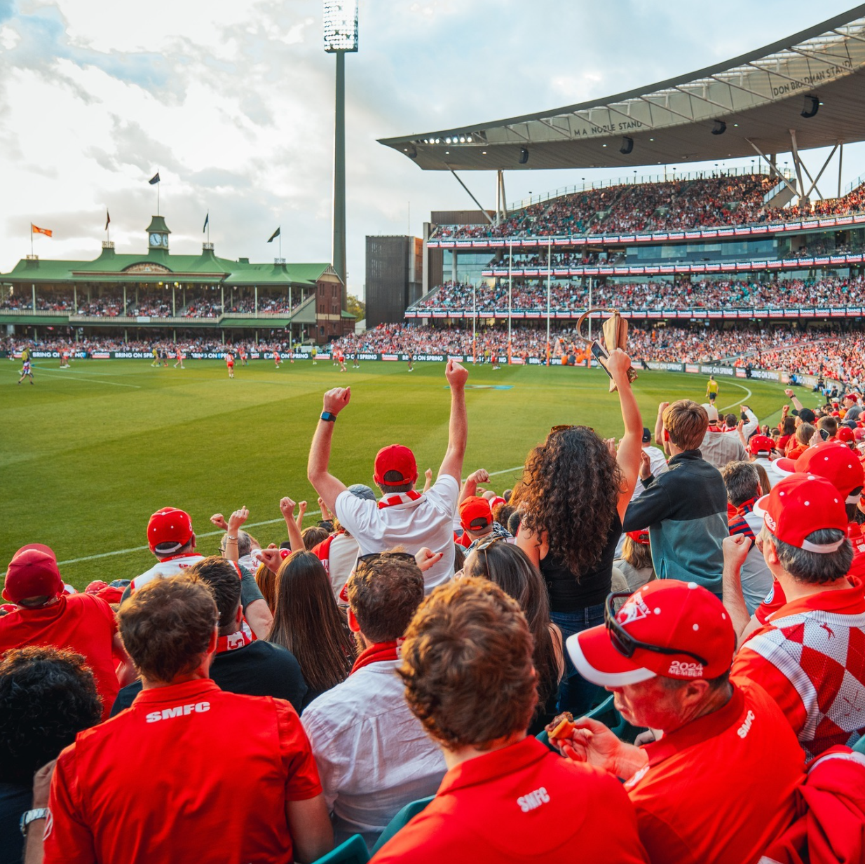 Crowd watching a sports match at a stadium with fans wearing red, some with scarves and hats, cheering and celebrating as the game goes on under partly cloudy skies.