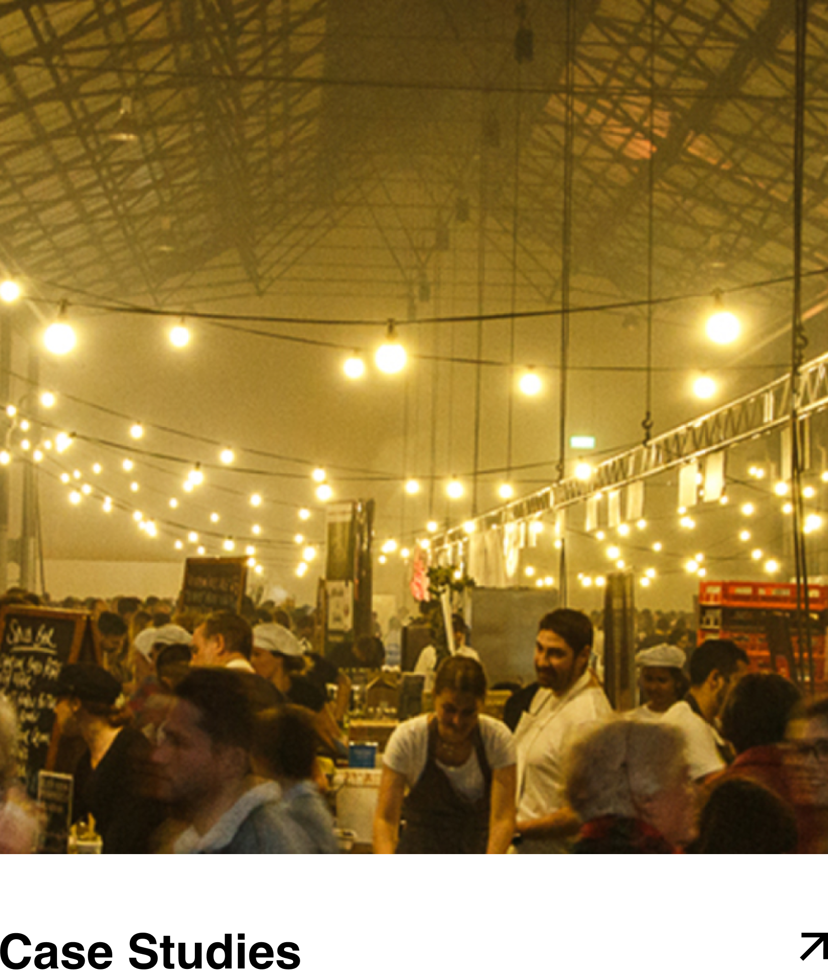 Indoor market or food festival with people and vendors, string lights overhead, and a large tent ceiling.