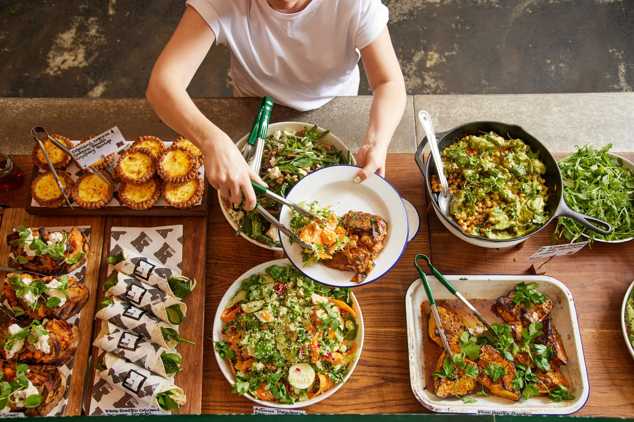 A person serves themselves from a table filled with various salads, grilled chicken, sliders, and sides at a buffet-style meal.