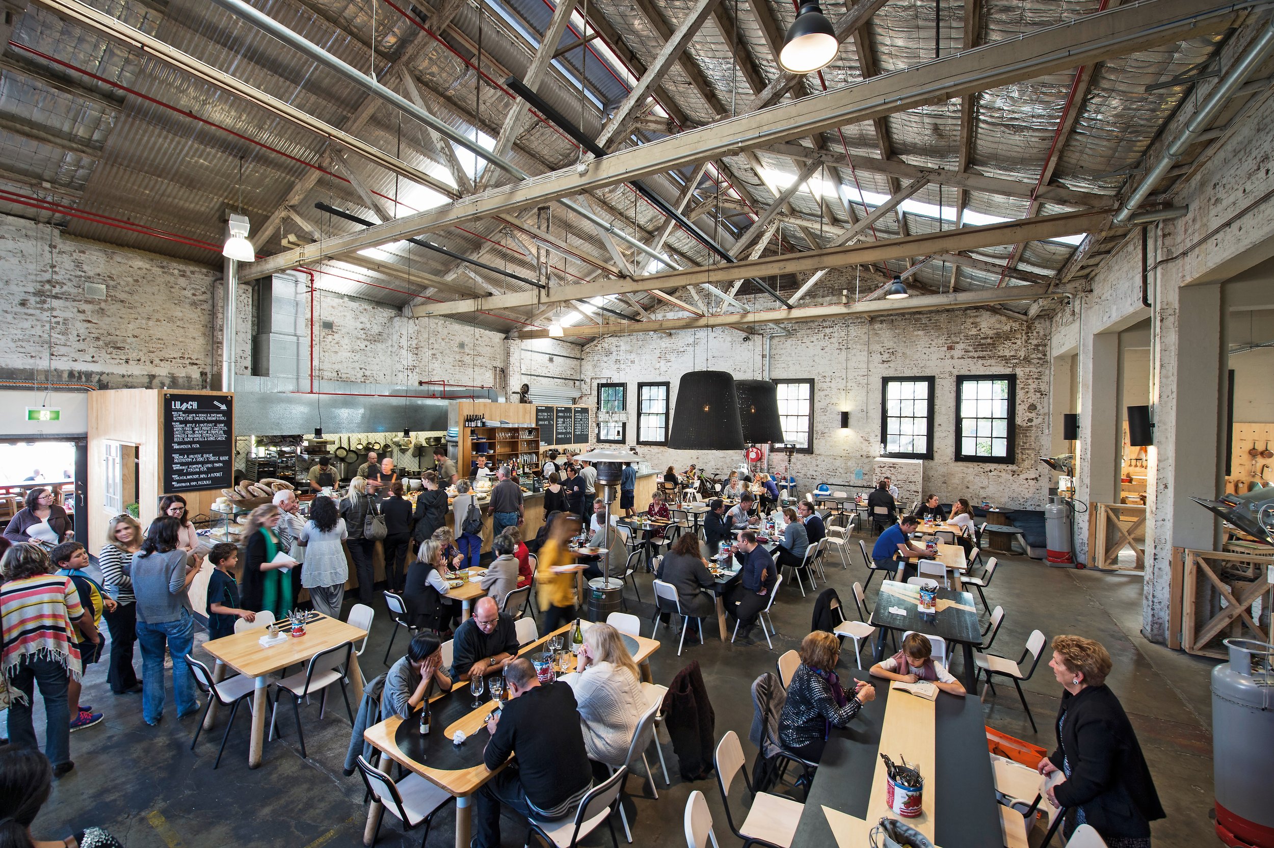 Interior of a busy industrial-style restaurant with high ceilings, exposed brick walls, and large windows. People are dining and ordering at the counter, with many tables occupied.