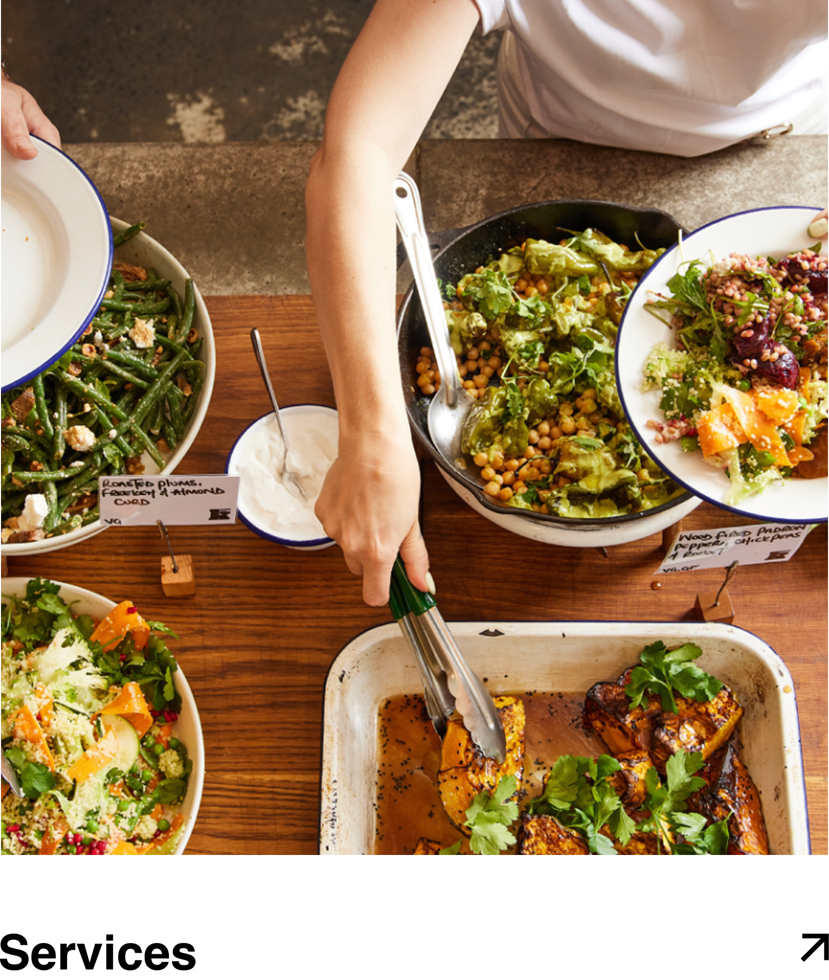 A person serving grilled chicken from a white baking dish onto a plate during a meal with various salads and side dishes on a wooden table.